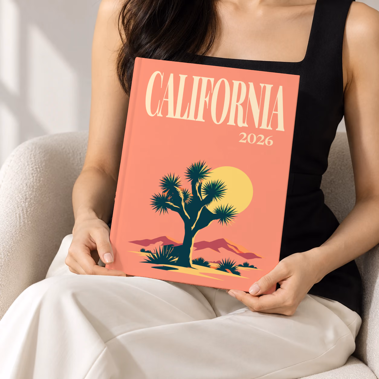 California hardcover photo book with the illustrated cover of a Joshua tree and desert hills under a low sun on coral, held upright in two hands by a woman in a black top, sitting in a cream boucle armchair