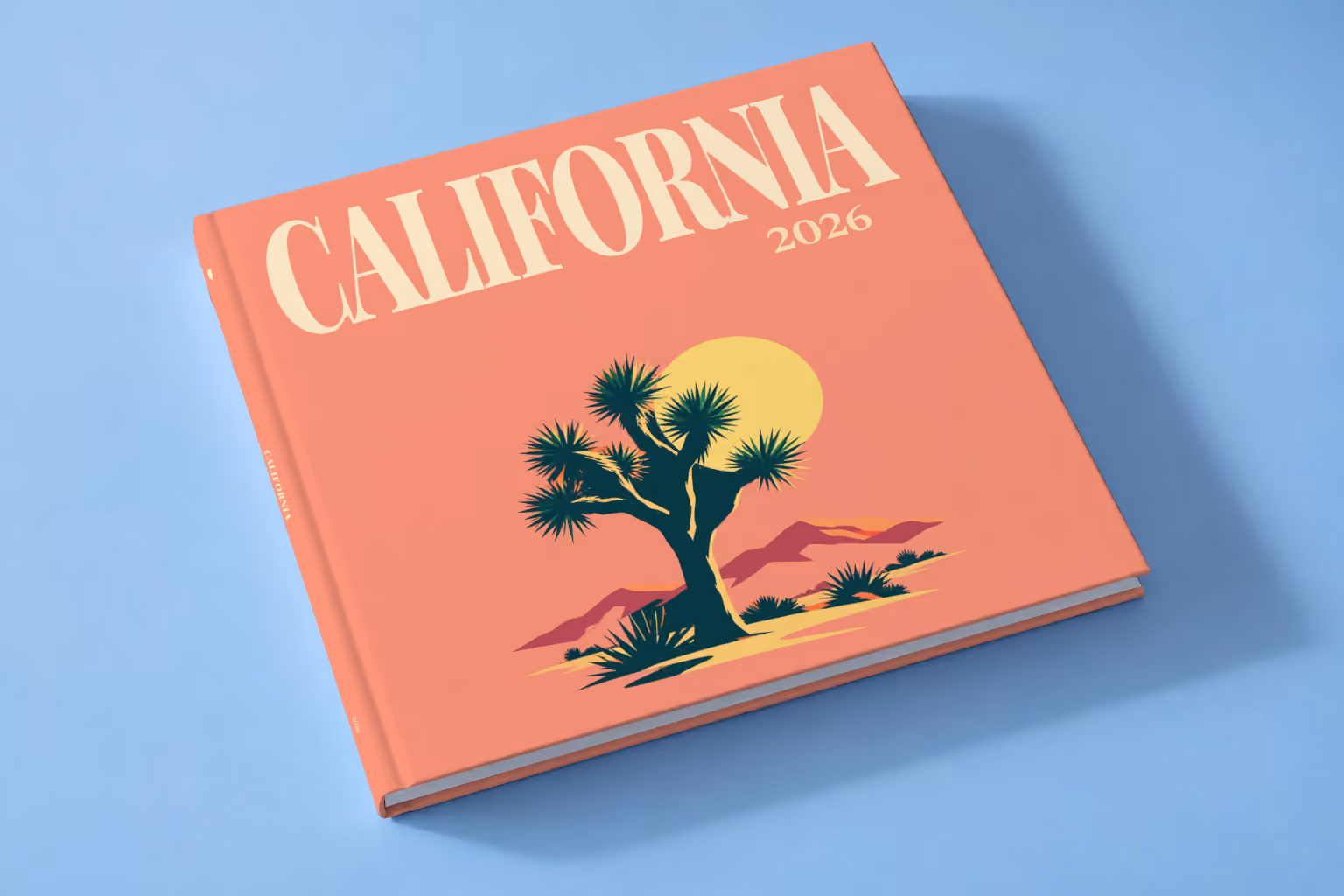 California square hardcover photo book with the illustrated cover of a Joshua tree and desert hills under a low sun on coral, lying on a pale blue studio backdrop