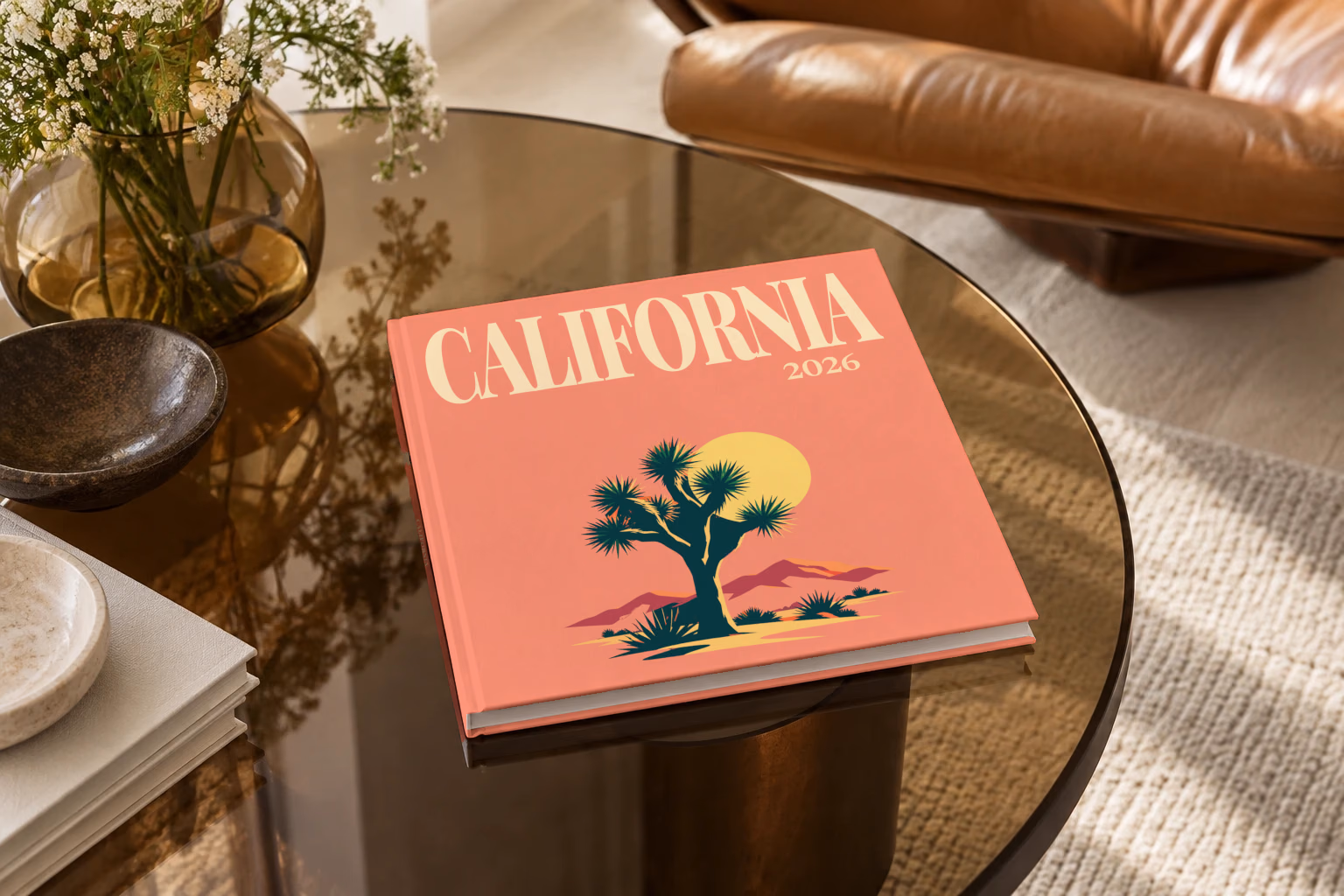 California square hardcover photo book with the illustrated cover of a Joshua tree and desert hills under a low sun on coral, on a smoked glass coffee table beside a leather armchair in warm afternoon light