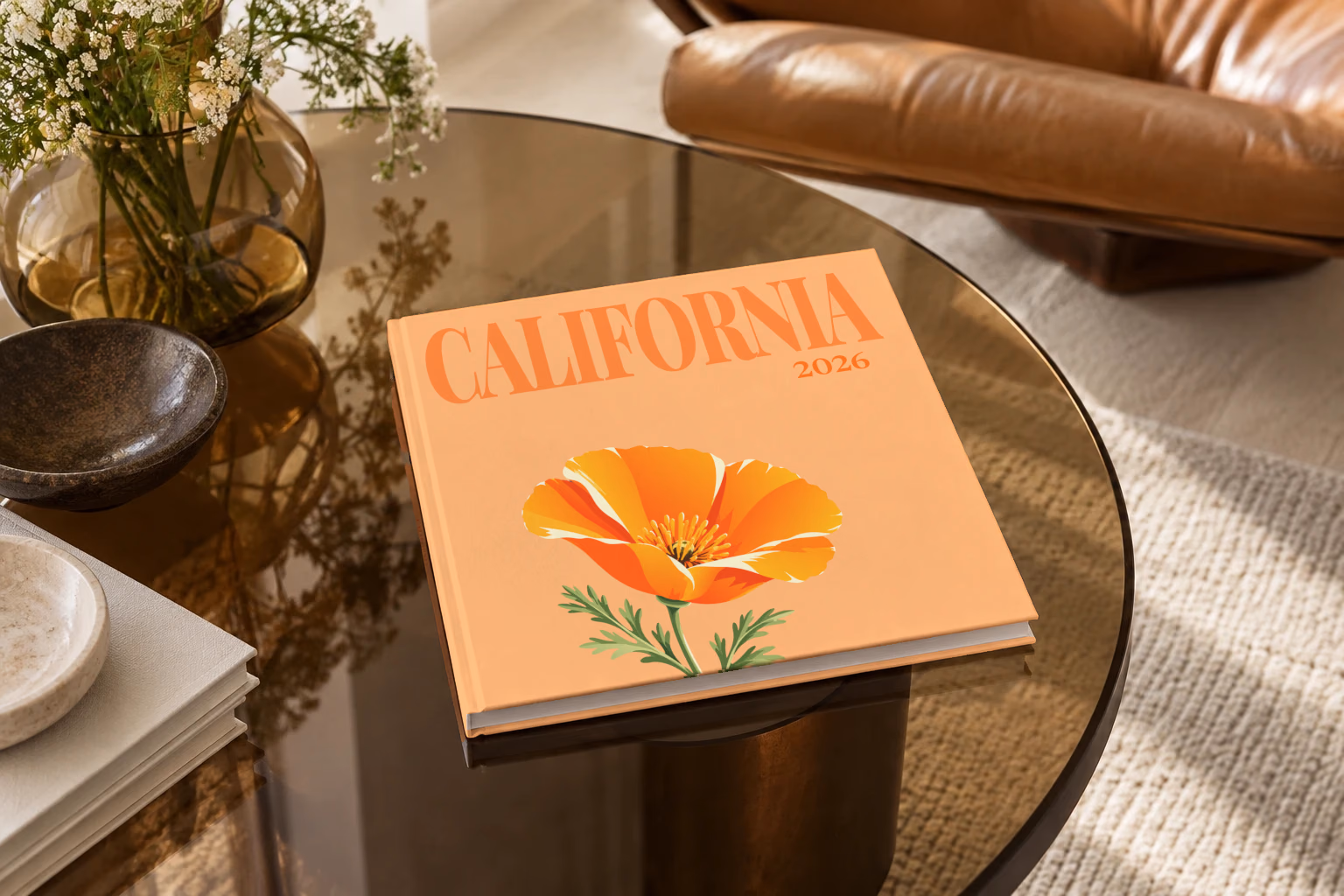 California square hardcover photo book with the illustrated cover of a California poppy on peach, on a smoked glass coffee table beside a leather armchair in warm afternoon light