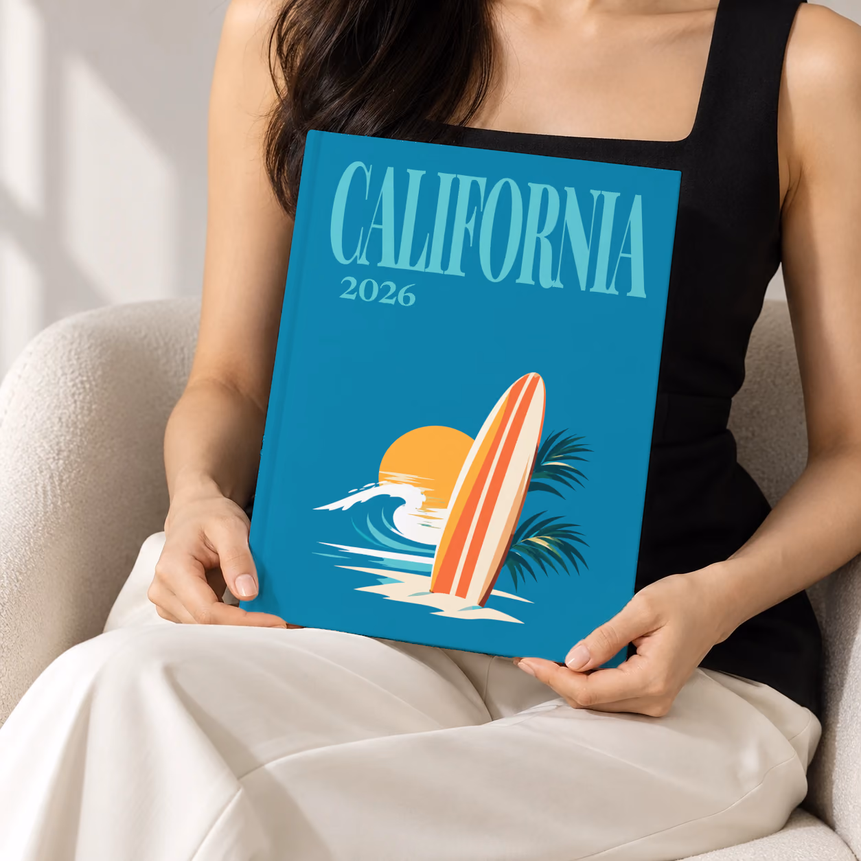 California hardcover photo book with the illustrated cover of a surfboard, a wave and the sun on teal, held upright in two hands by a woman in a black top, sitting in a cream boucle armchair