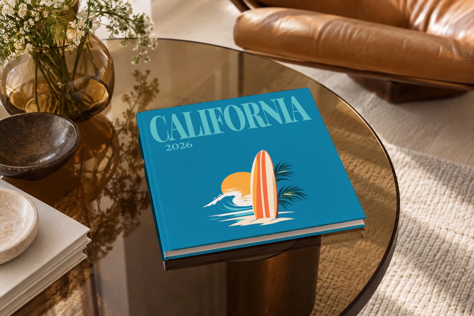 California square hardcover photo book with the illustrated cover of a surfboard, a wave and the sun on teal, on a smoked glass coffee table beside a leather armchair in warm afternoon light