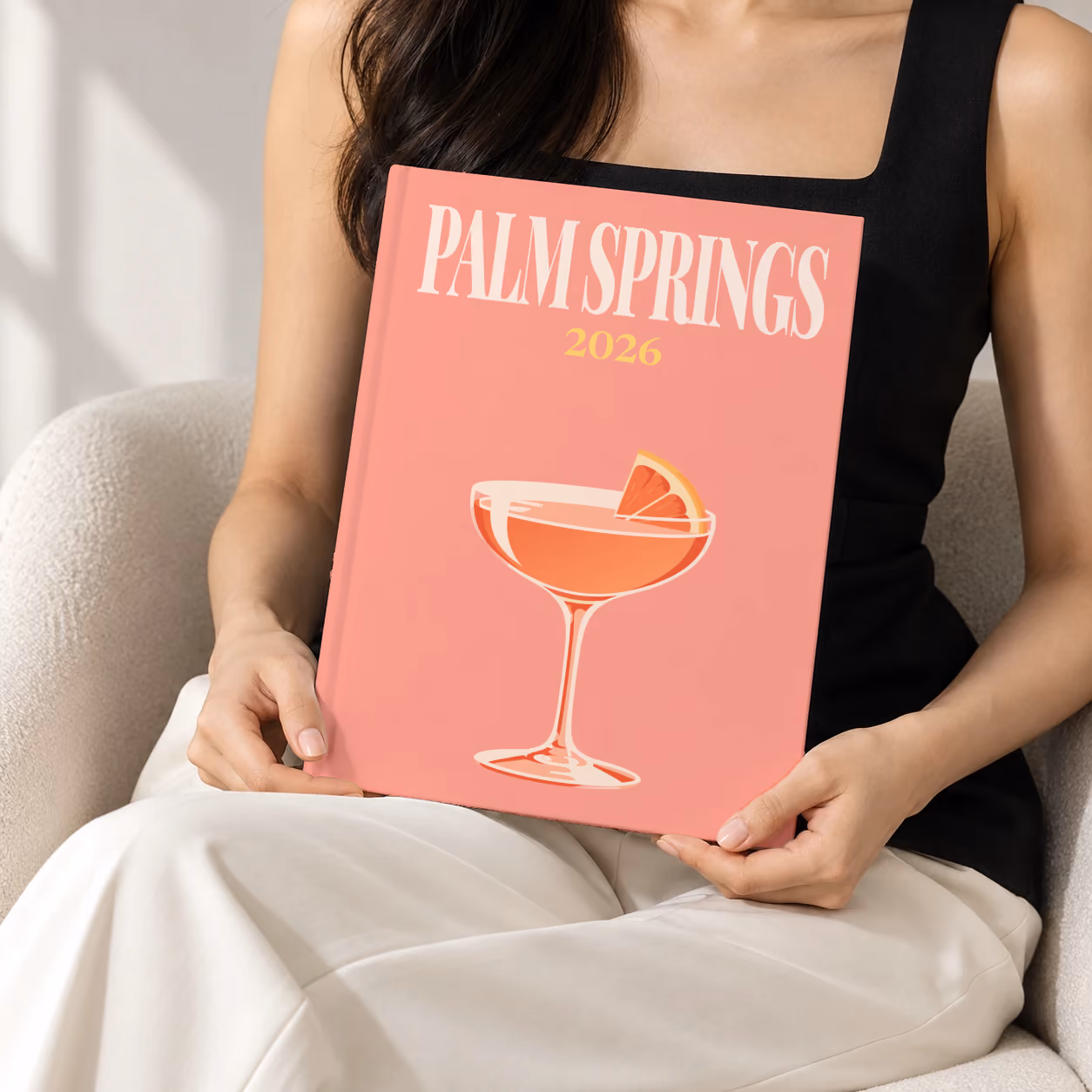 California hardcover photo book with the Palm Springs illustrated cover of a coupe cocktail with grapefruit on salmon pink, held upright in two hands by a woman in a black top, sitting in a cream boucle armchair