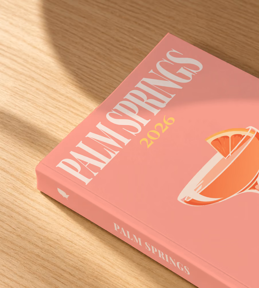 California softcover photo book with the Palm Springs illustrated cover of a coupe cocktail with grapefruit on salmon pink, lying on an oak desk in dappled afternoon sunlight