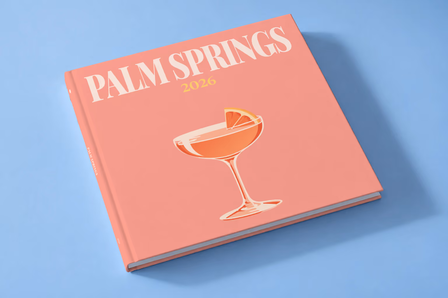 California square hardcover photo book with the Palm Springs illustrated cover of a coupe cocktail with grapefruit on salmon pink, lying on a pale blue studio backdrop