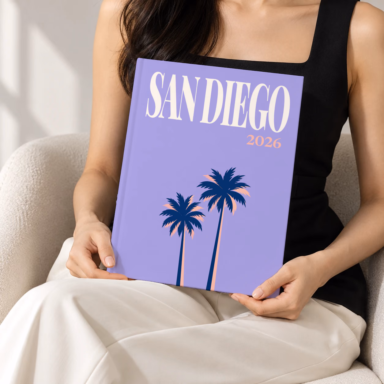 California hardcover photo book with the San Diego illustrated cover of palm trees on lilac, held upright in two hands by a woman in a black top, sitting in a cream boucle armchair