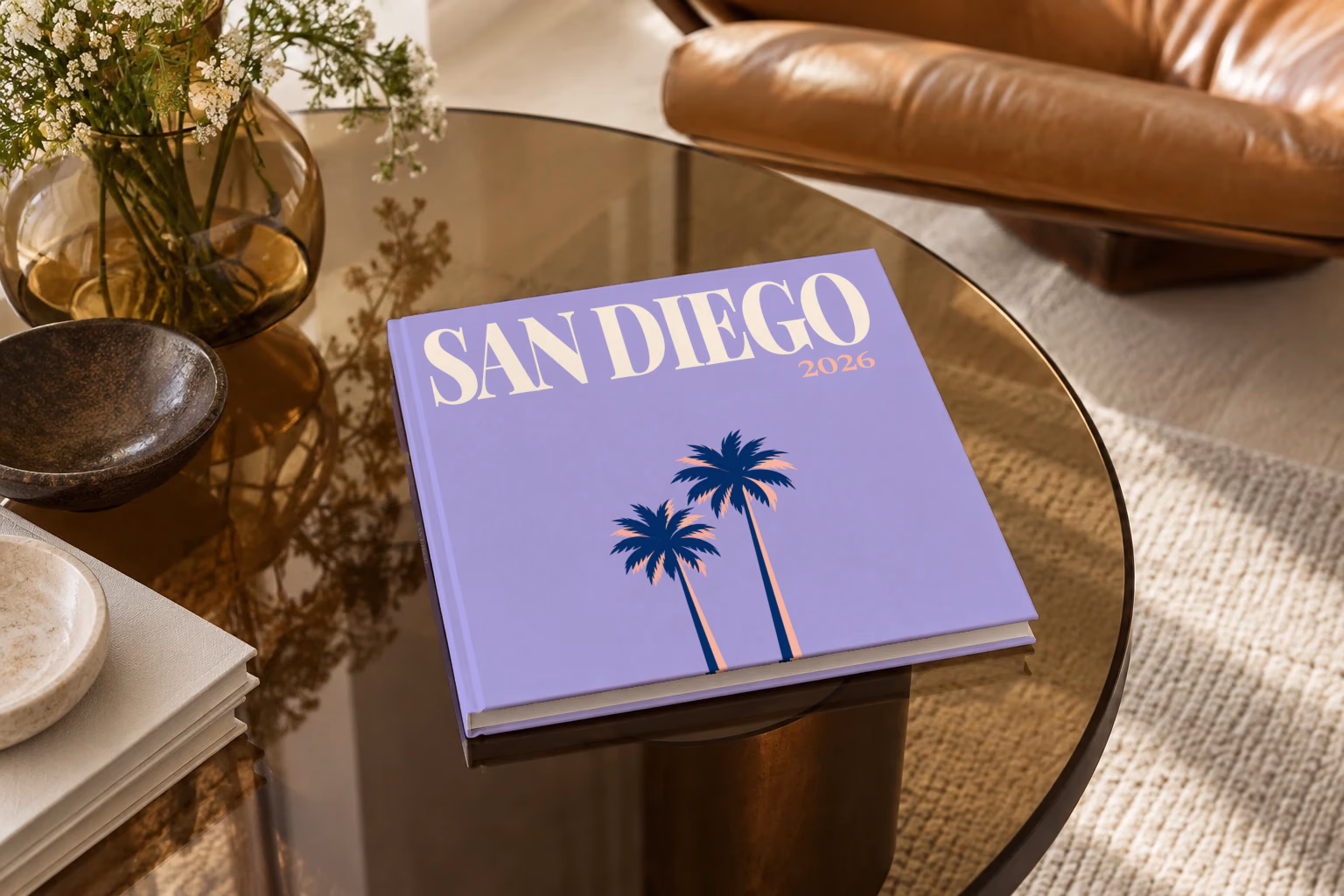 California square hardcover photo book with the San Diego illustrated cover of palm trees on lilac, on a smoked glass coffee table beside a leather armchair in warm afternoon light