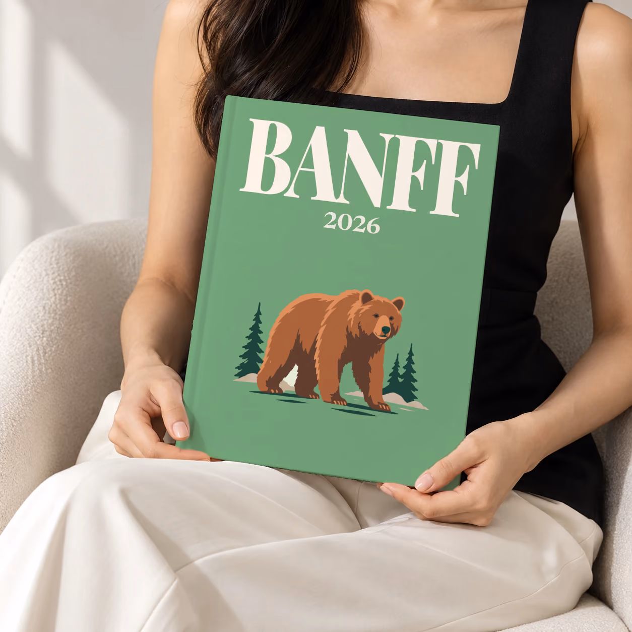 Canada hardcover photo book with the Banff illustrated cover of a grizzly bear and pines on sage green, held upright in two hands by a woman in a black top, sitting in a cream boucle armchair