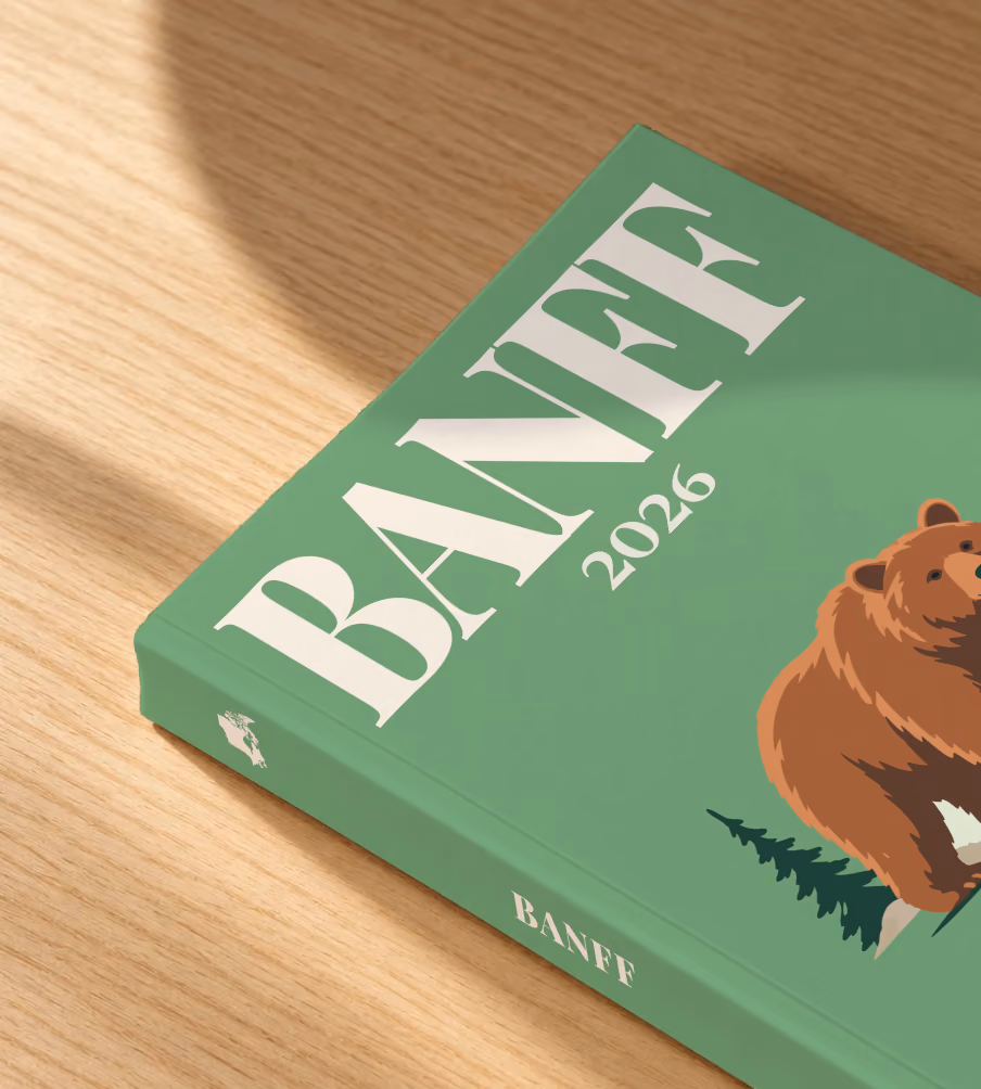 Canada softcover photo book with the Banff illustrated cover of a grizzly bear and pines on sage green, lying on an oak desk in dappled afternoon sunlight