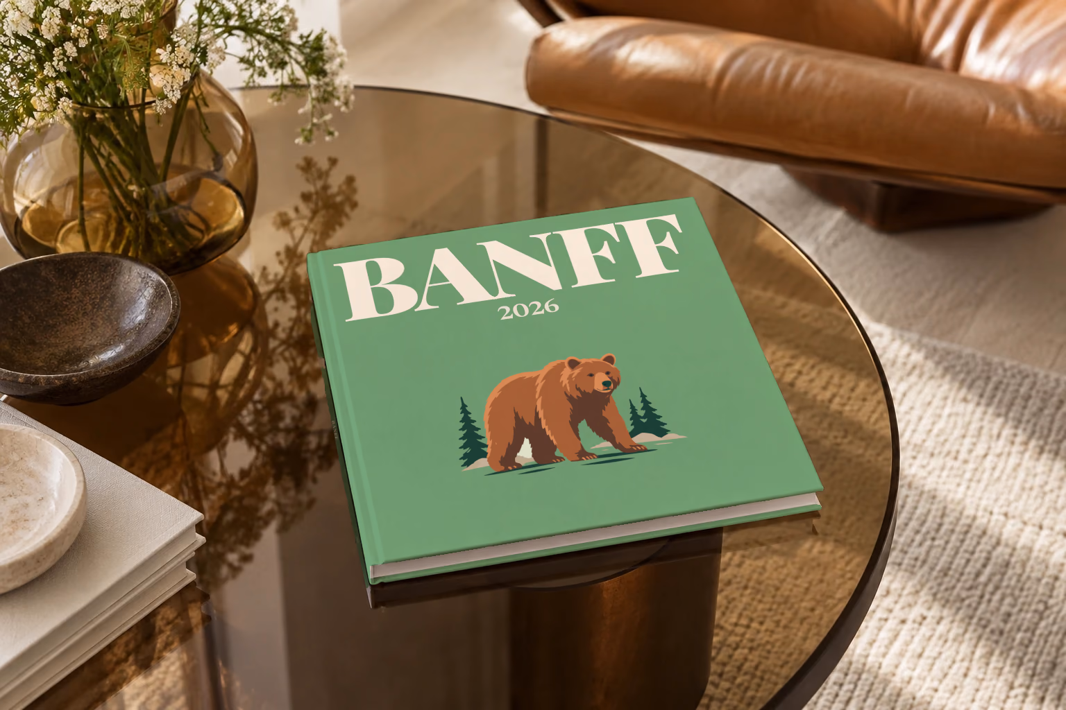 Canada square hardcover photo book with the Banff illustrated cover of a grizzly bear and pines on sage green, on a smoked glass coffee table beside a leather armchair in warm afternoon light