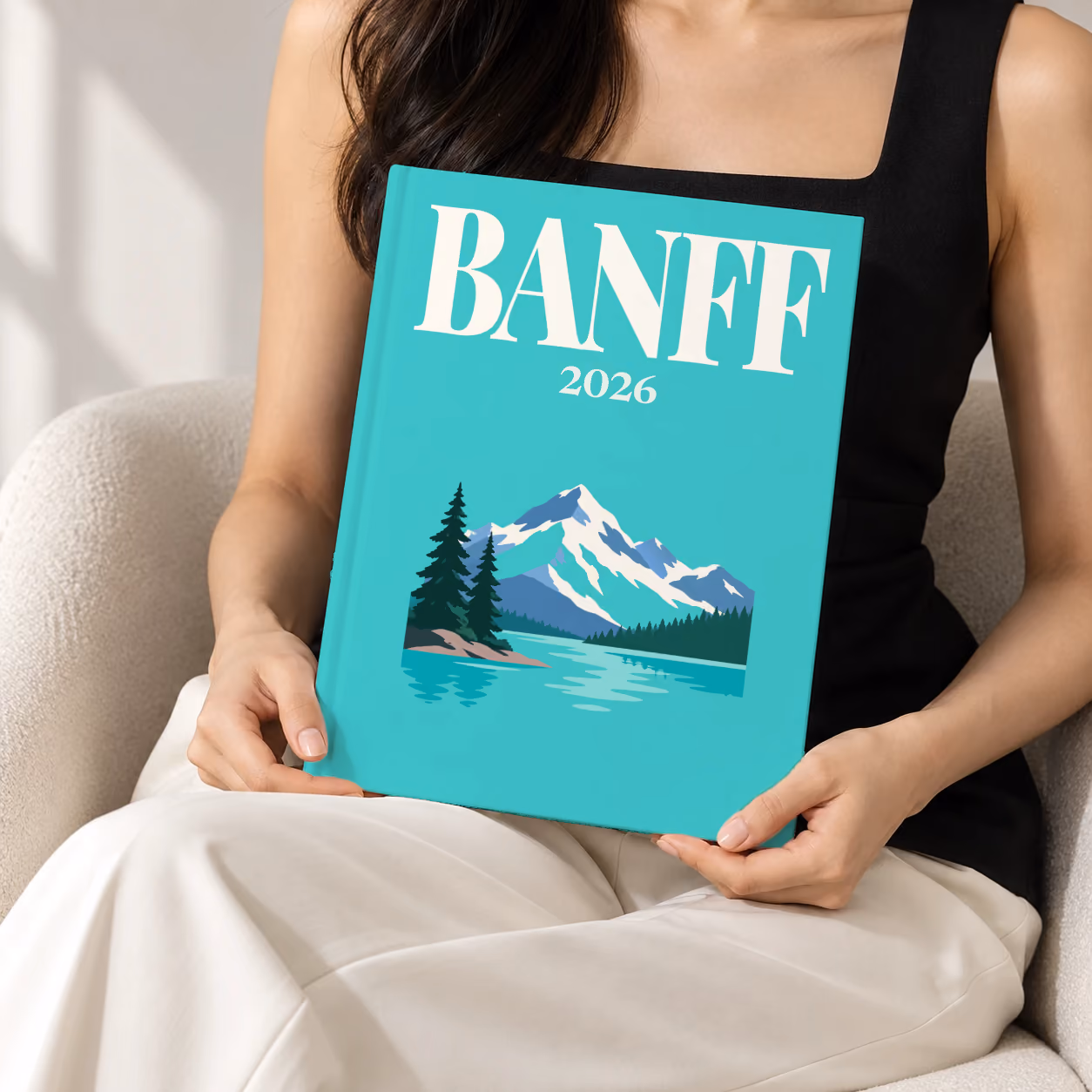 Canada hardcover photo book with the Banff illustrated cover of a mountain lake, snowy peaks and pines on turquoise, held upright in two hands by a woman in a black top, sitting in a cream boucle armchair