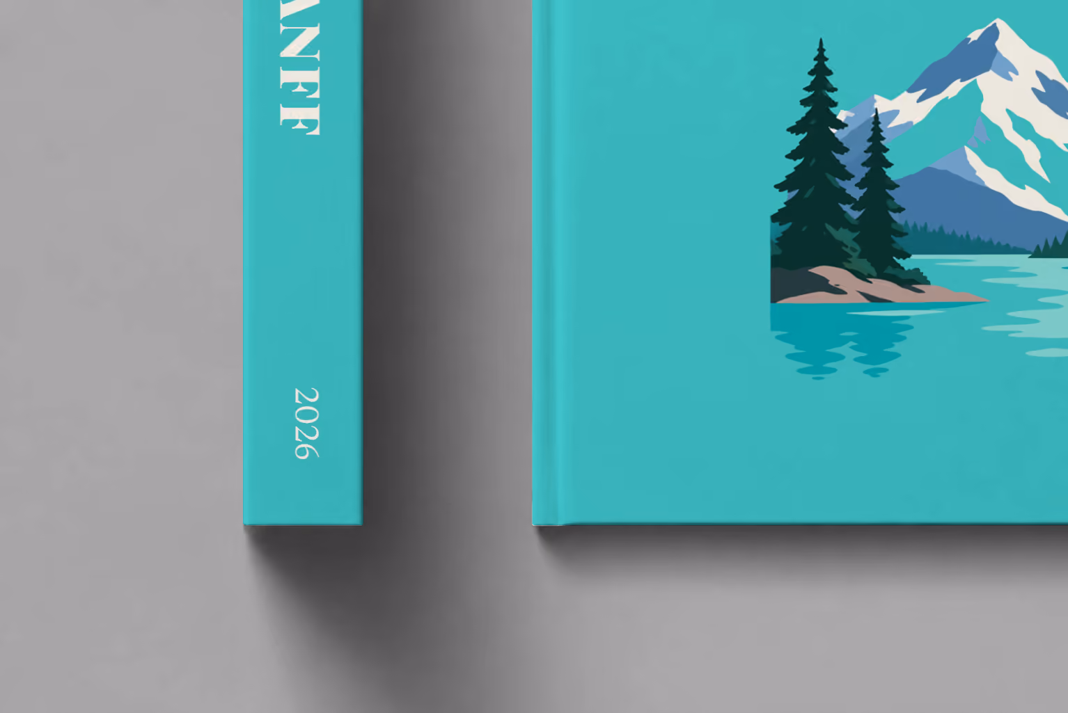 Canada hardcover photo book with the Banff illustrated cover of a mountain lake, snowy peaks and pines on turquoise, shown close up beside its spine on a pale grey studio surface