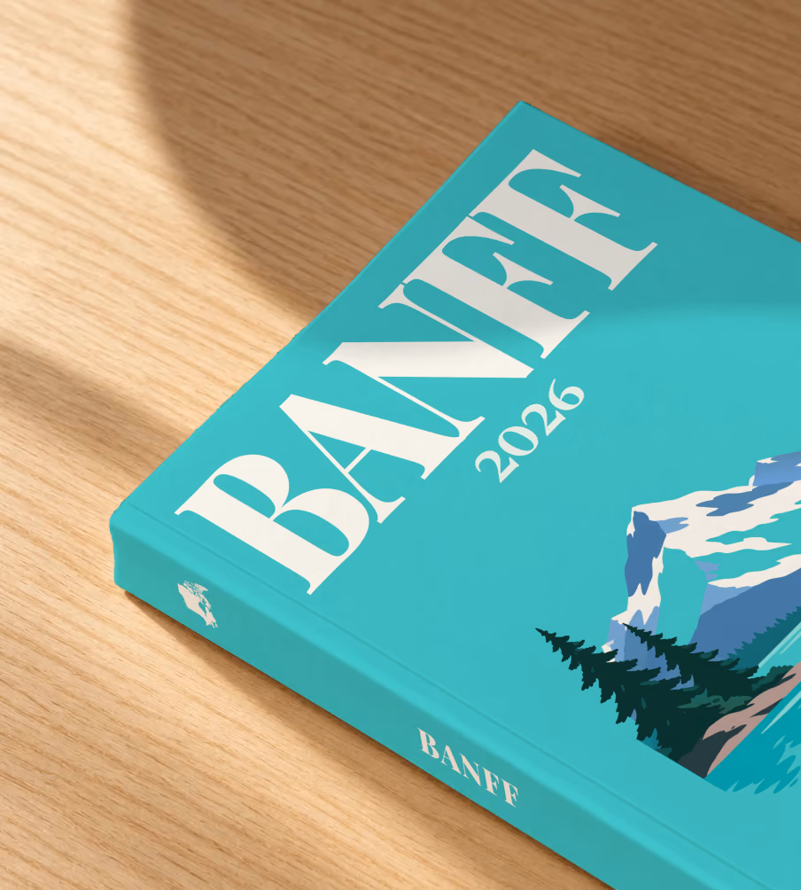 Canada softcover photo book with the Banff illustrated cover of a mountain lake, snowy peaks and pines on turquoise, lying on an oak desk in dappled afternoon sunlight
