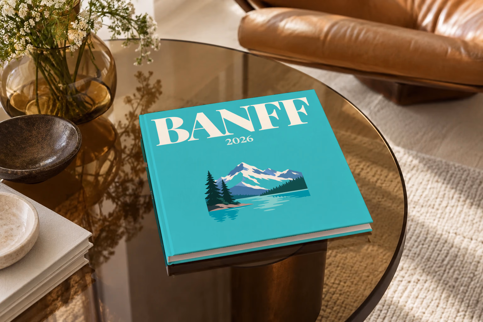 Canada square hardcover photo book with the Banff illustrated cover of a mountain lake, snowy peaks and pines on turquoise, on a smoked glass coffee table beside a leather armchair in warm afternoon light