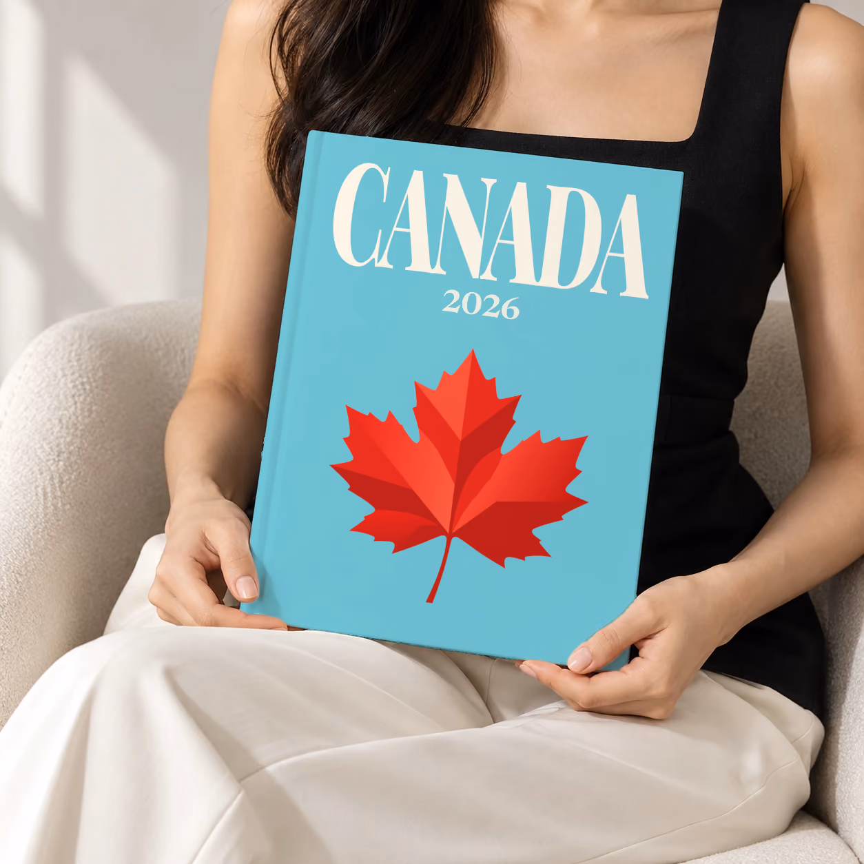 Canada hardcover photo book with the illustrated cover of a red maple leaf on turquoise, held upright in two hands by a woman in a black top, sitting in a cream boucle armchair