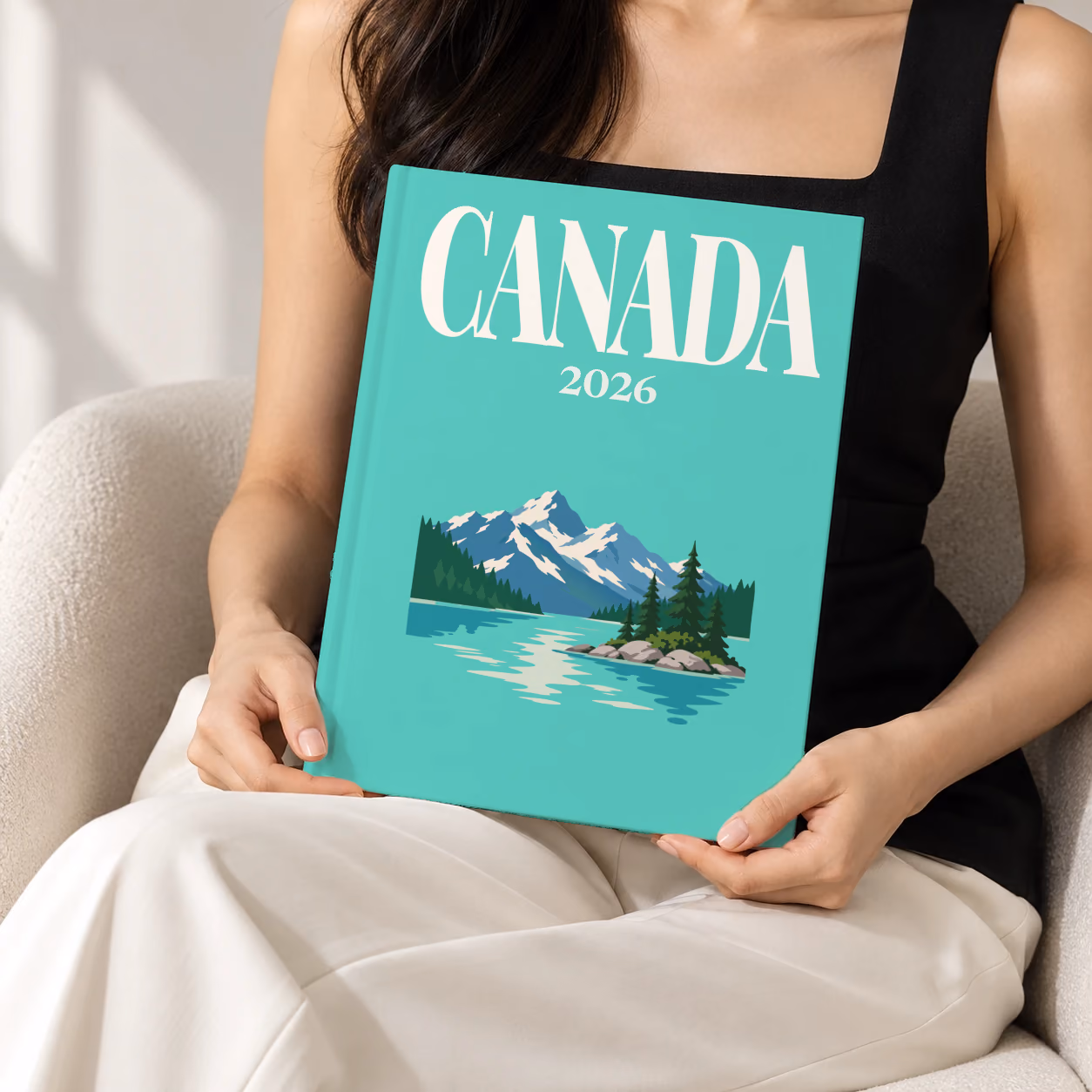 Canada hardcover photo book with the illustrated cover of a mountain lake with a pine-covered islet on mint green, held upright in two hands by a woman in a black top, sitting in a cream boucle armchair