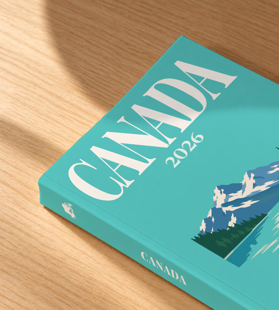 Canada softcover photo book with the illustrated cover of a mountain lake with a pine-covered islet on mint green, lying on an oak desk in dappled afternoon sunlight