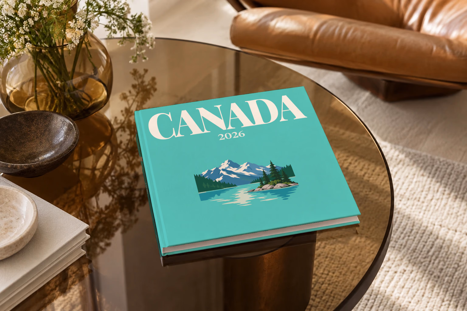 Canada square hardcover photo book with the illustrated cover of a mountain lake with a pine-covered islet on mint green, on a smoked glass coffee table beside a leather armchair in warm afternoon light