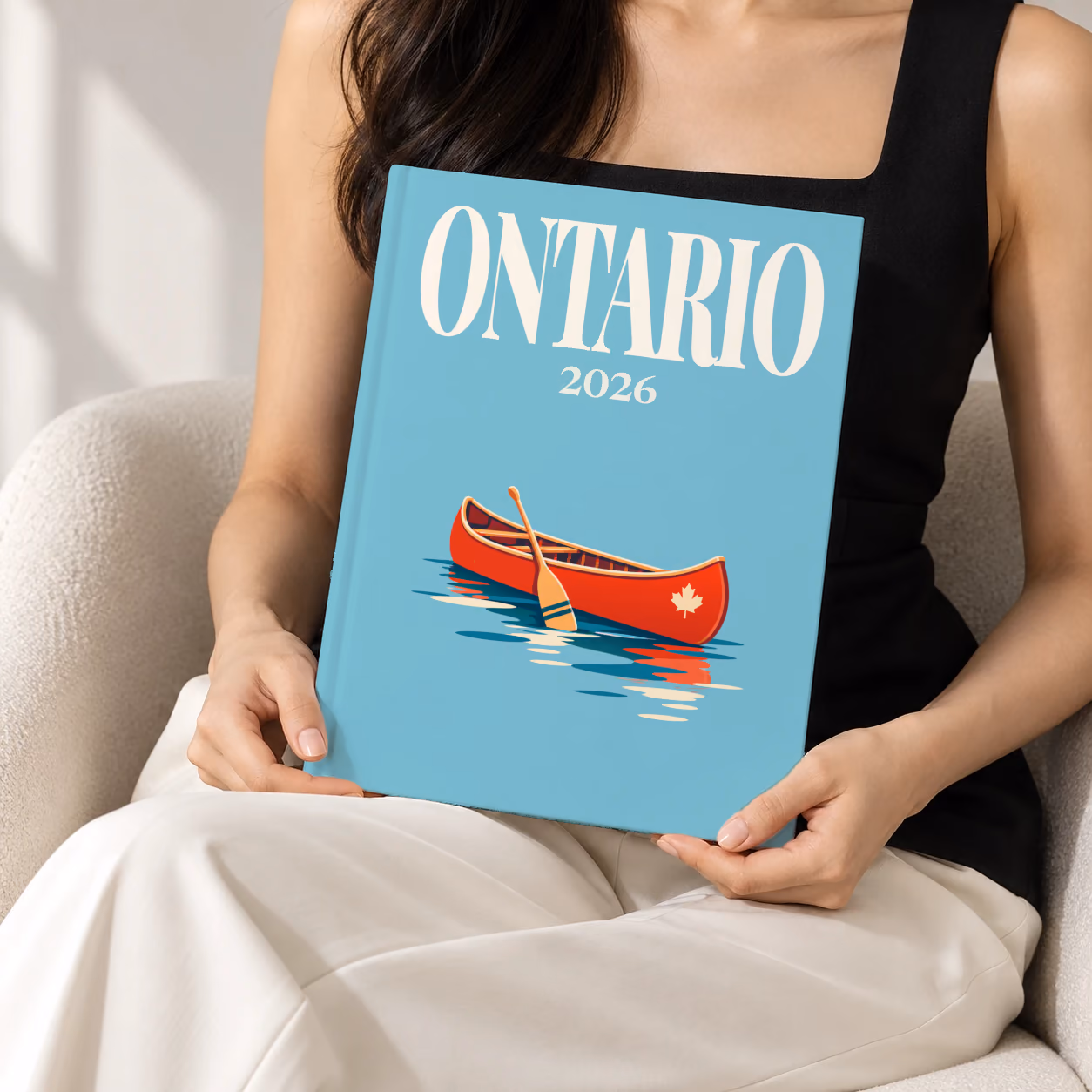 Canada hardcover photo book with the Ontario illustrated cover of a red canoe on a lake on dusty blue, held upright in two hands by a woman in a black top, sitting in a cream boucle armchair
