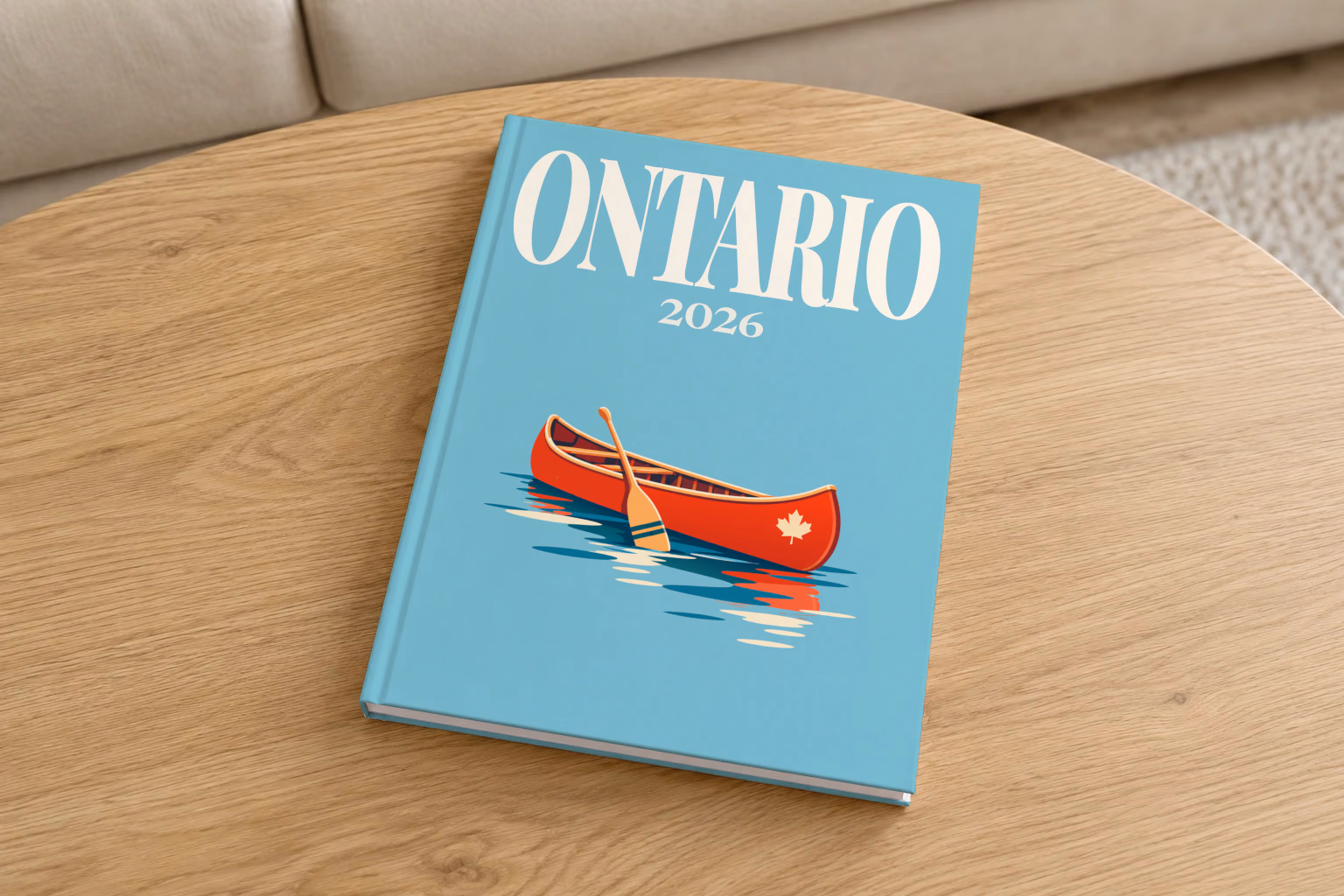 Canada hardcover photo book with the Ontario illustrated cover of a red canoe on a lake on dusty blue, on a round oak coffee table in a bright living room
