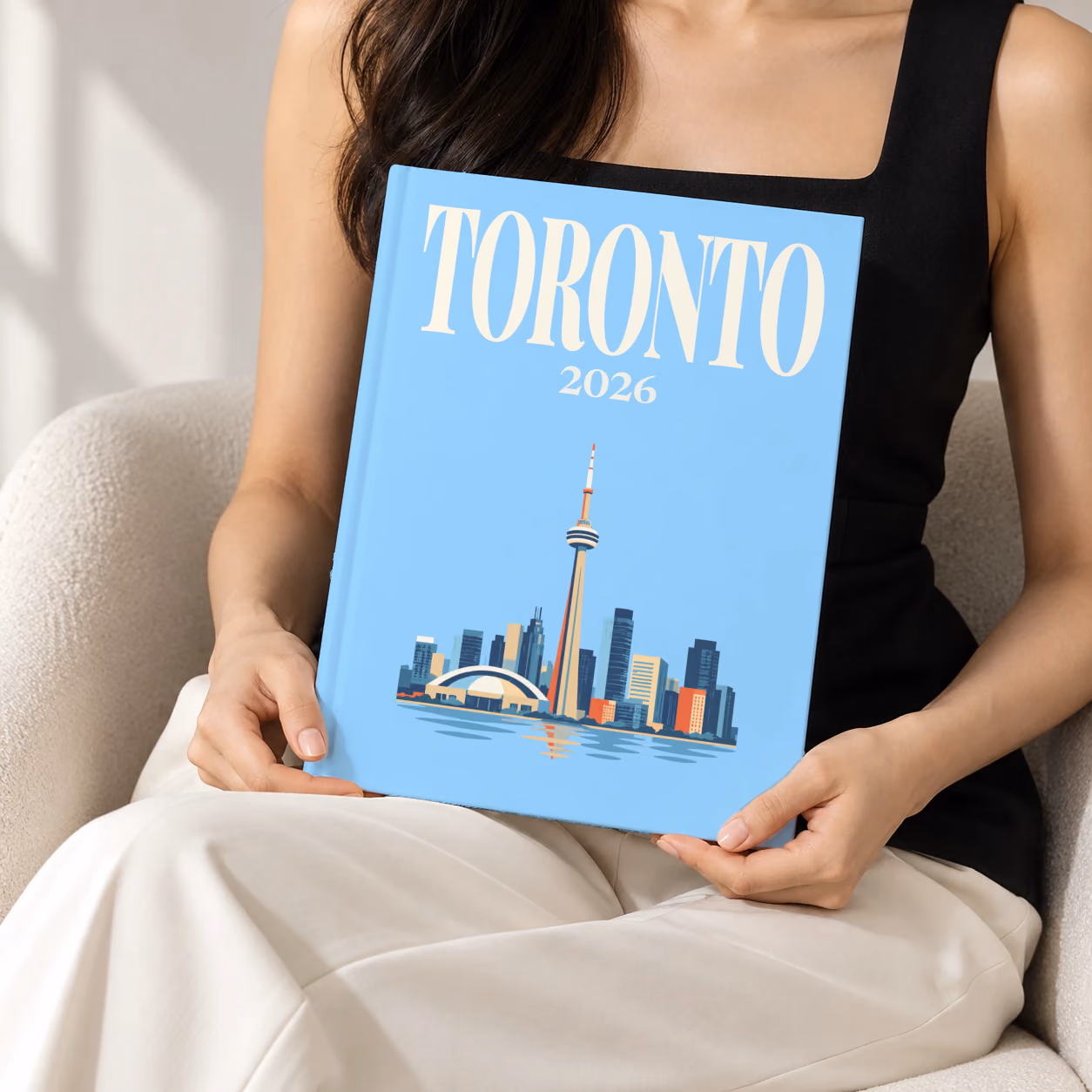 Canada hardcover photo book with the Toronto illustrated cover of the Toronto skyline and CN Tower on pale blue, held upright in two hands by a woman in a black top, sitting in a cream boucle armchair