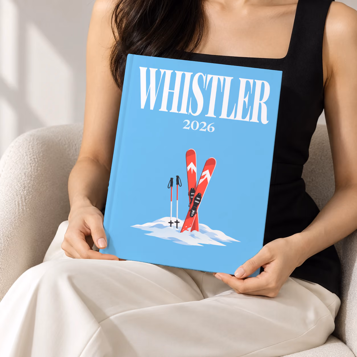 Canada hardcover photo book with the Whistler illustrated cover of red skis and poles in snow on sky blue, held upright in two hands by a woman in a black top, sitting in a cream boucle armchair