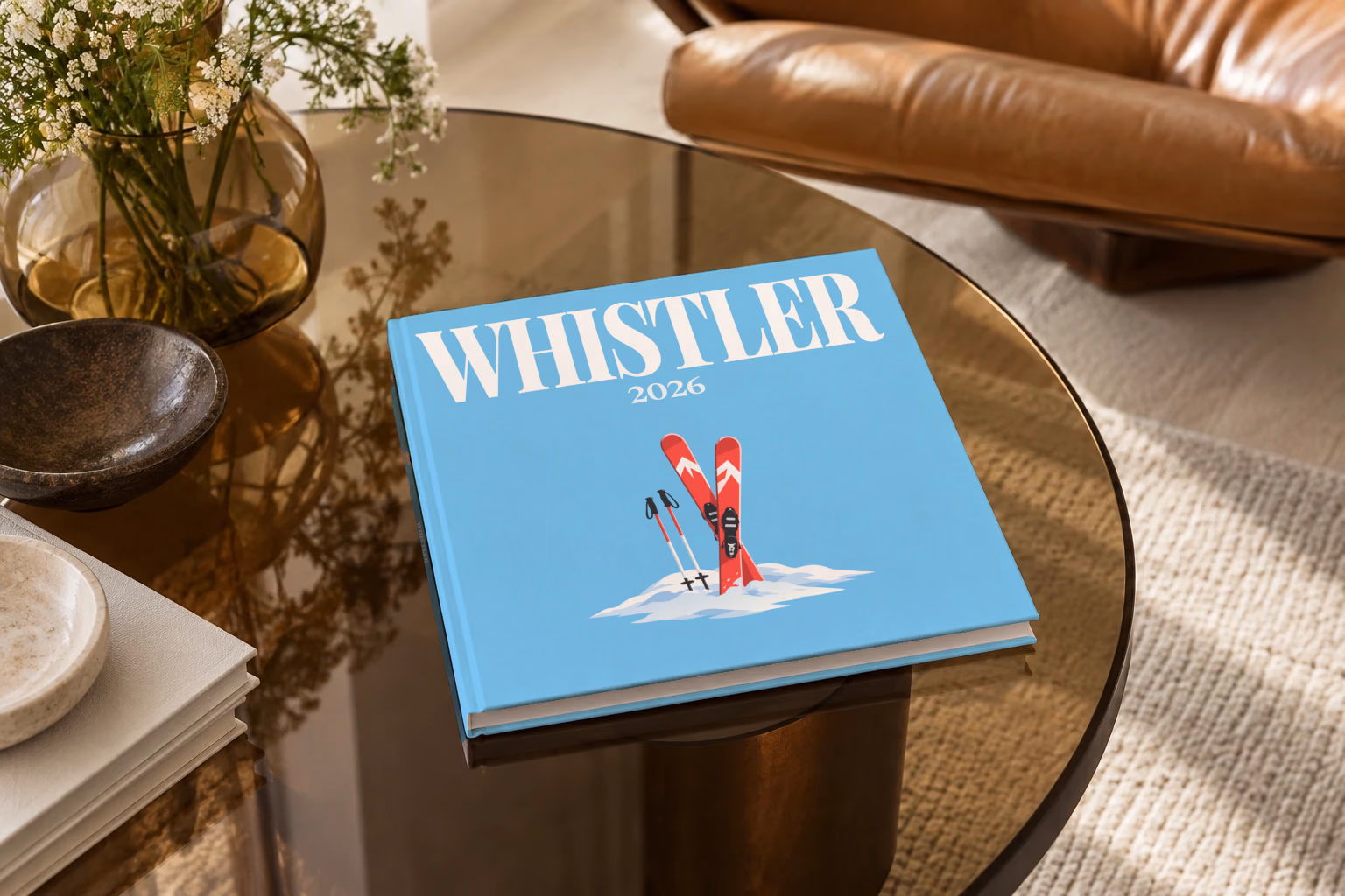Canada square hardcover photo book with the Whistler illustrated cover of red skis and poles in snow on sky blue, on a smoked glass coffee table beside a leather armchair in warm afternoon light