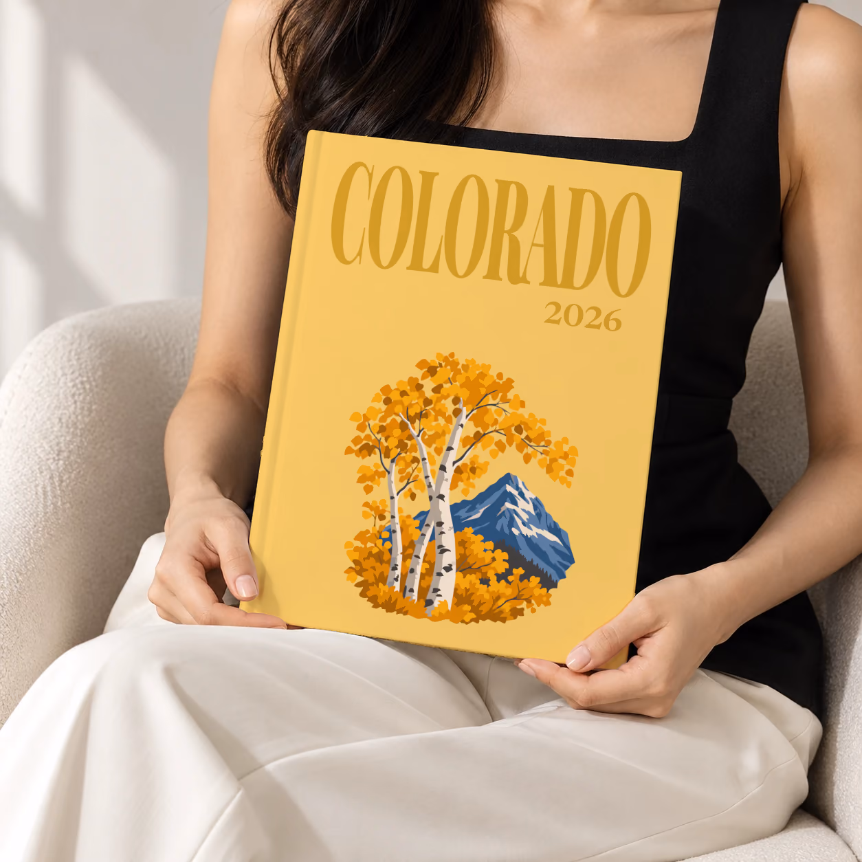 Colorado hardcover photo book with the illustrated cover of golden aspen trees and a mountain peak on mustard yellow, held upright in two hands by a woman in a black top, sitting in a cream boucle armchair