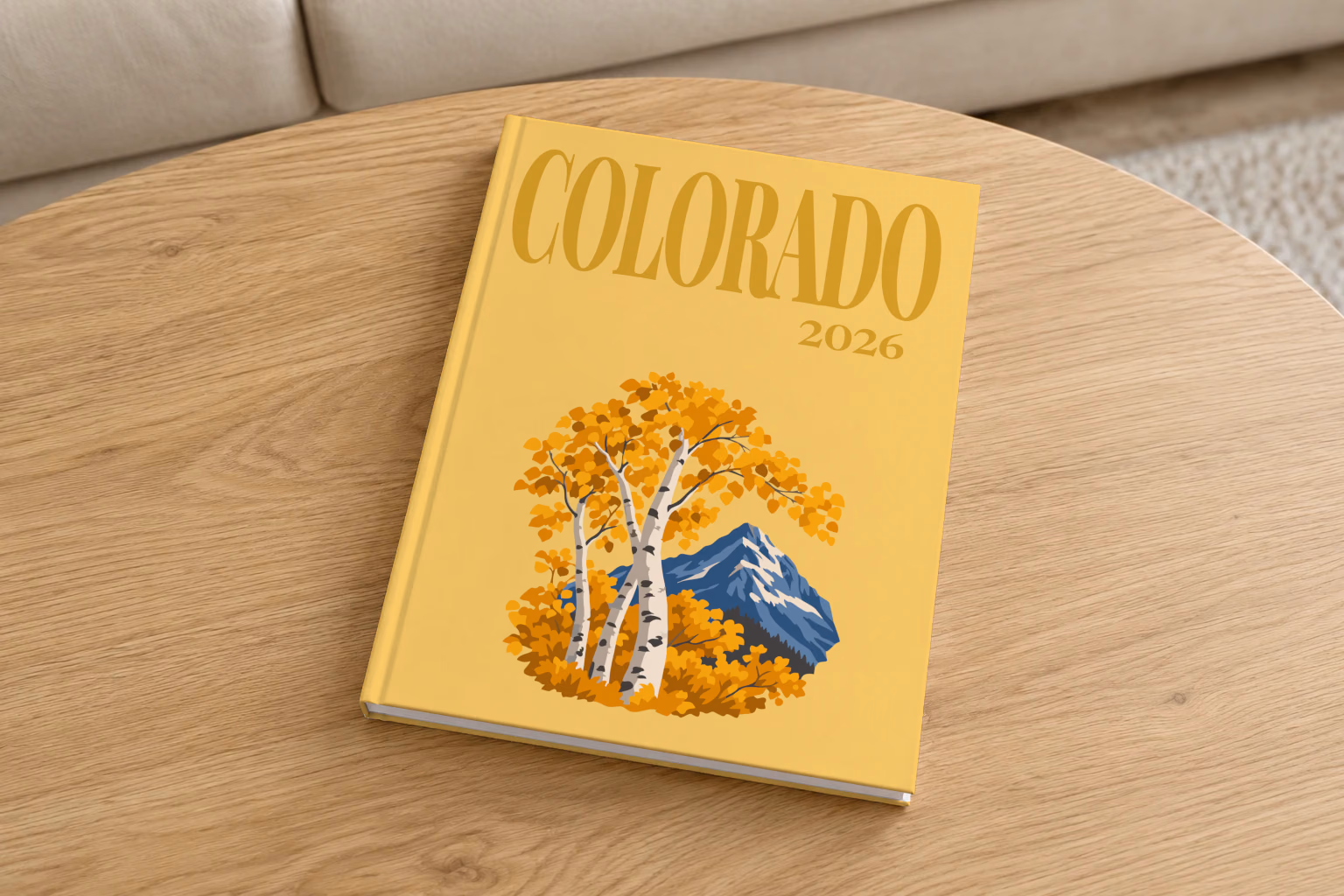 Colorado hardcover photo book with the illustrated cover of golden aspen trees and a mountain peak on mustard yellow, on a round oak coffee table in a bright living room