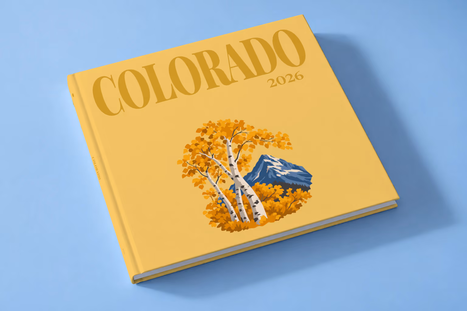 Colorado square hardcover photo book with the illustrated cover of golden aspen trees and a mountain peak on mustard yellow, lying on a pale blue studio backdrop