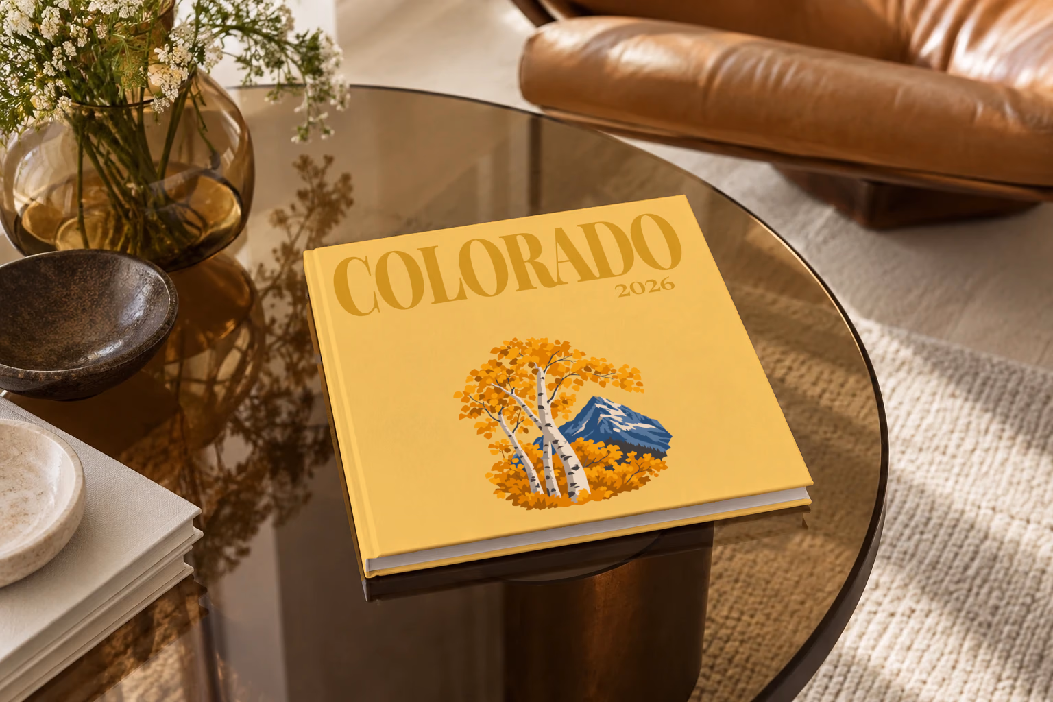 Colorado square hardcover photo book with the illustrated cover of golden aspen trees and a mountain peak on mustard yellow, on a smoked glass coffee table beside a leather armchair in warm afternoon light