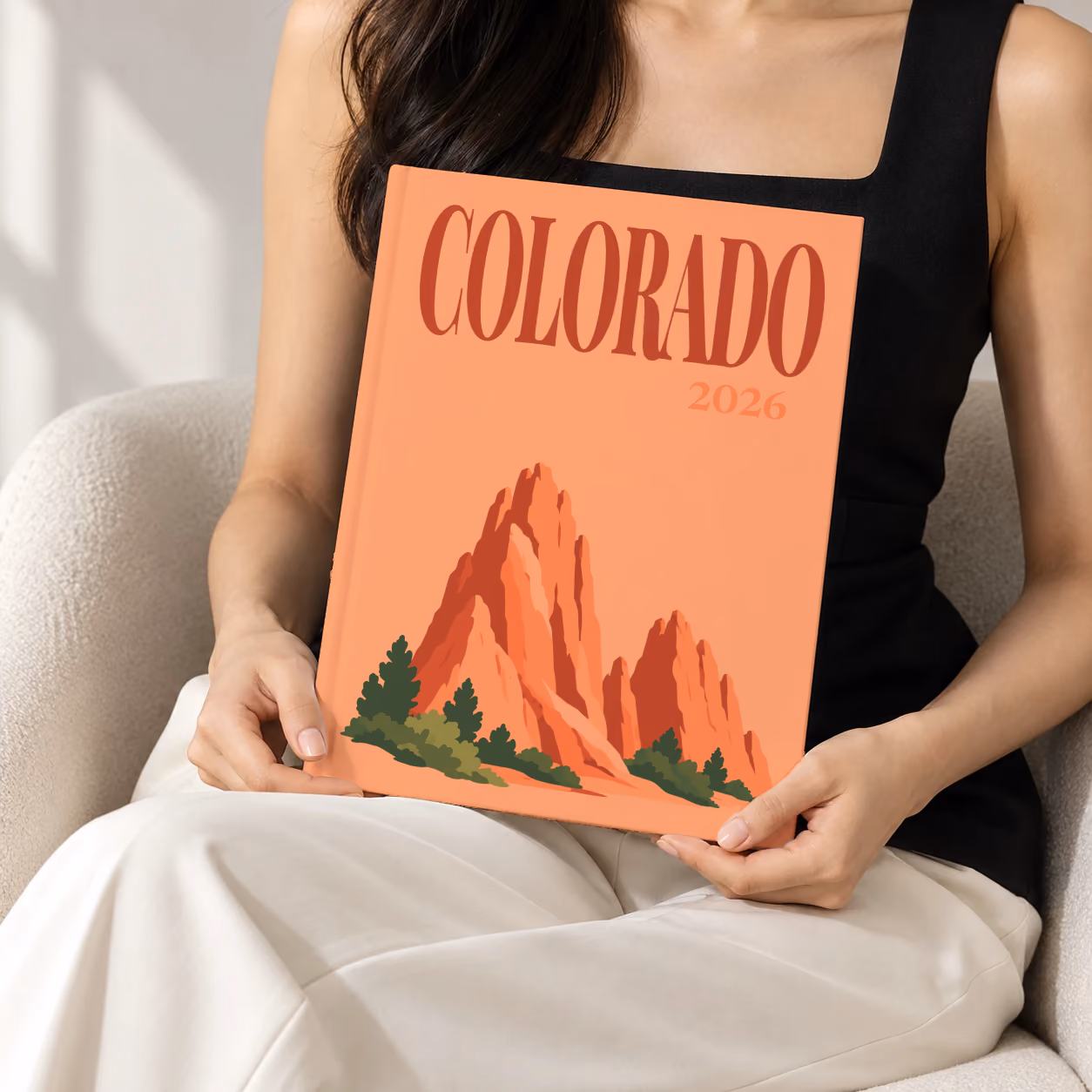 Colorado hardcover photo book with the illustrated cover of the red rock formations of Garden of the Gods on peach, held upright in two hands by a woman in a black top, sitting in a cream boucle armchair