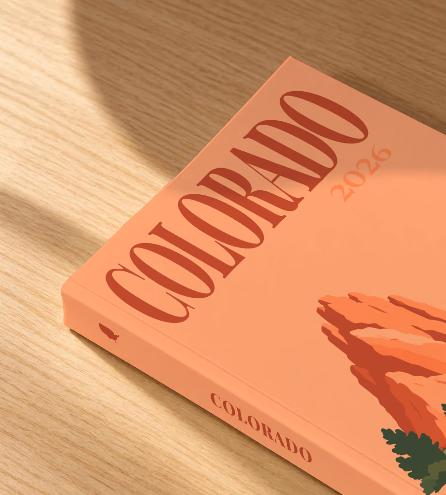 Colorado softcover photo book with the illustrated cover of the red rock formations of Garden of the Gods on peach, lying on an oak desk in dappled afternoon sunlight