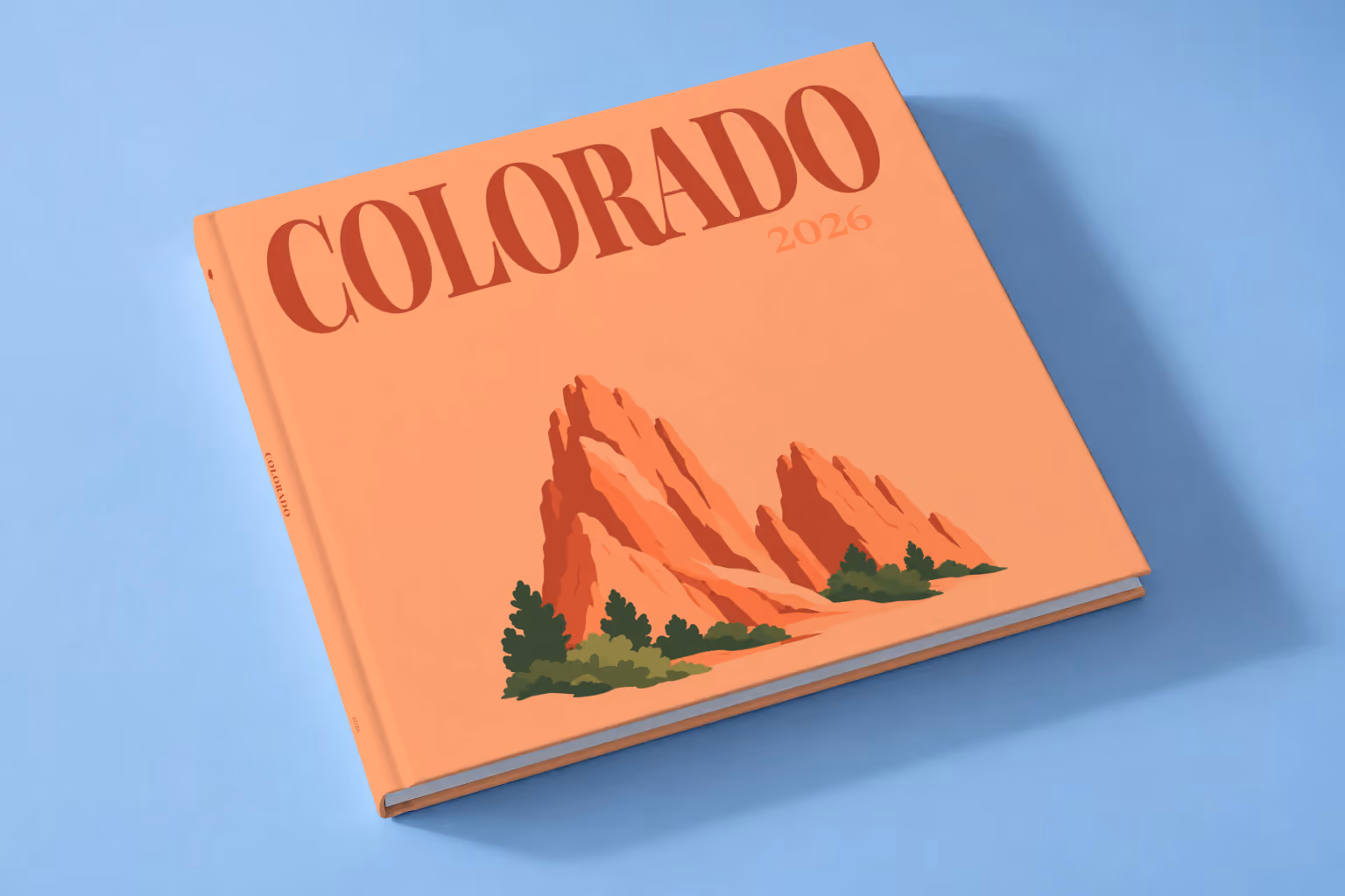 Colorado square hardcover photo book with the illustrated cover of the red rock formations of Garden of the Gods on peach, lying on a pale blue studio backdrop