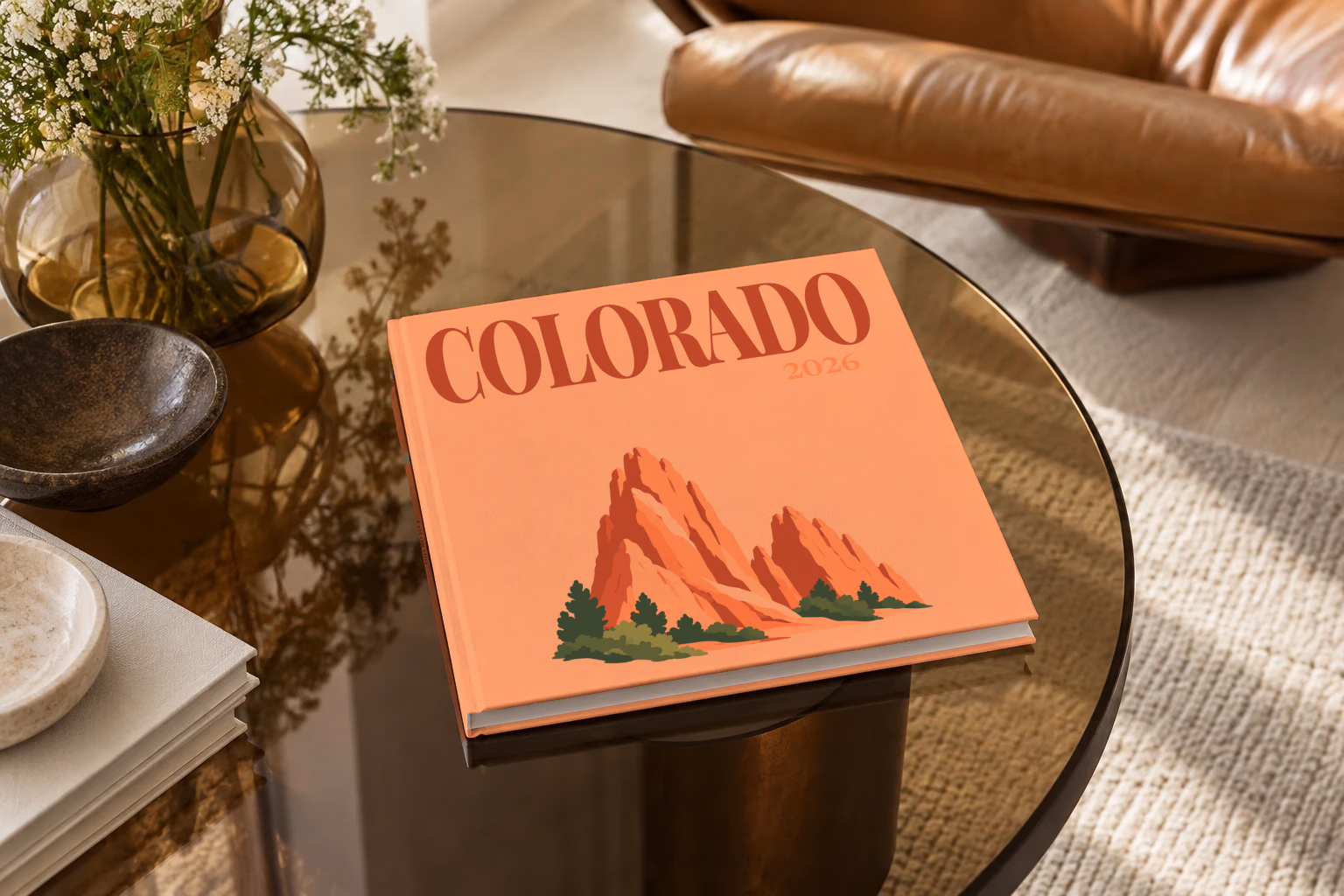 Colorado square hardcover photo book with the illustrated cover of the red rock formations of Garden of the Gods on peach, on a smoked glass coffee table beside a leather armchair in warm afternoon light