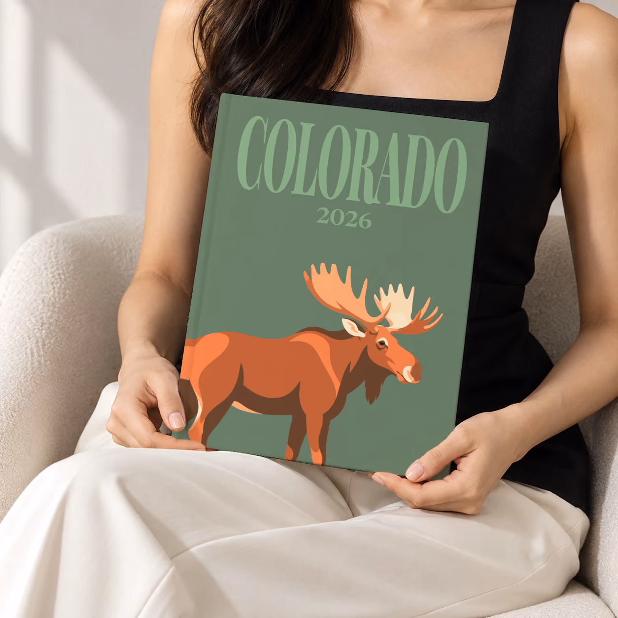 Colorado hardcover photo book with the illustrated cover of a bull moose on sage green, held upright in two hands by a woman in a black top, sitting in a cream boucle armchair