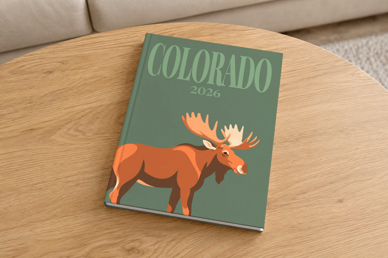 Colorado hardcover photo book with the illustrated cover of a bull moose on sage green, on a round oak coffee table in a bright living room