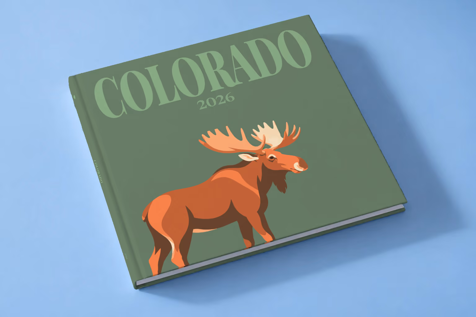 Colorado square hardcover photo book with the illustrated cover of a bull moose on sage green, lying on a pale blue studio backdrop