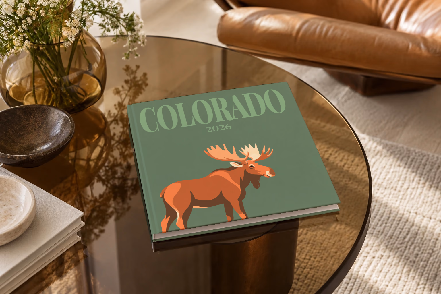 Colorado square hardcover photo book with the illustrated cover of a bull moose on sage green, on a smoked glass coffee table beside a leather armchair in warm afternoon light