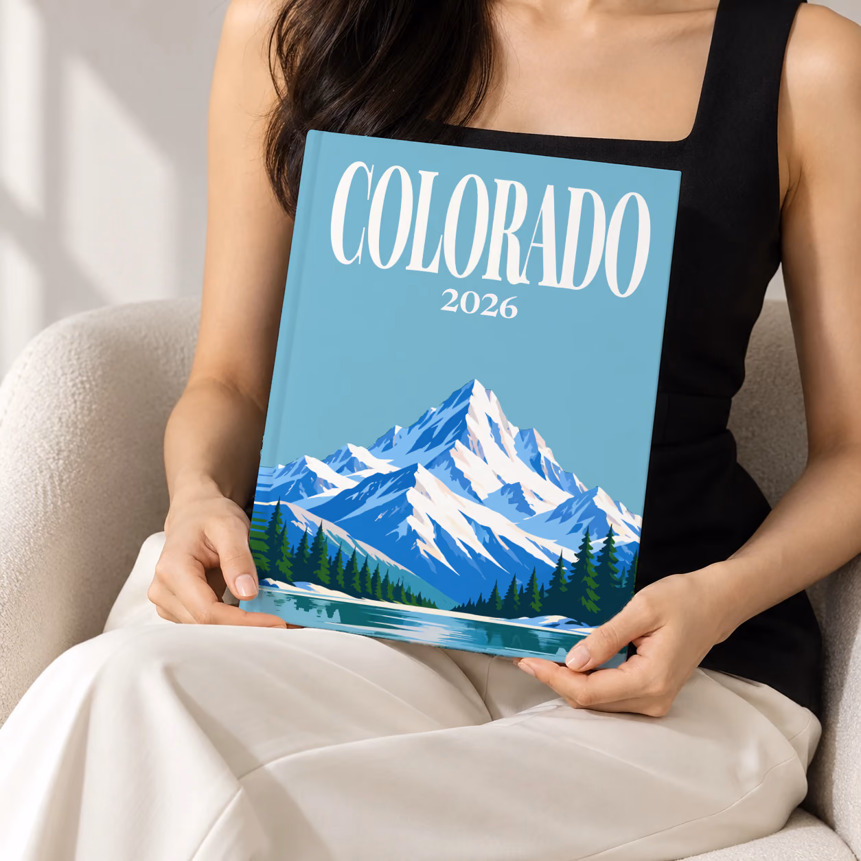 Colorado hardcover photo book with the illustrated cover of a snow-capped Rocky Mountain peak above a pine-lined lake on teal blue, held upright in two hands by a woman in a black top, sitting in a cream boucle armchair