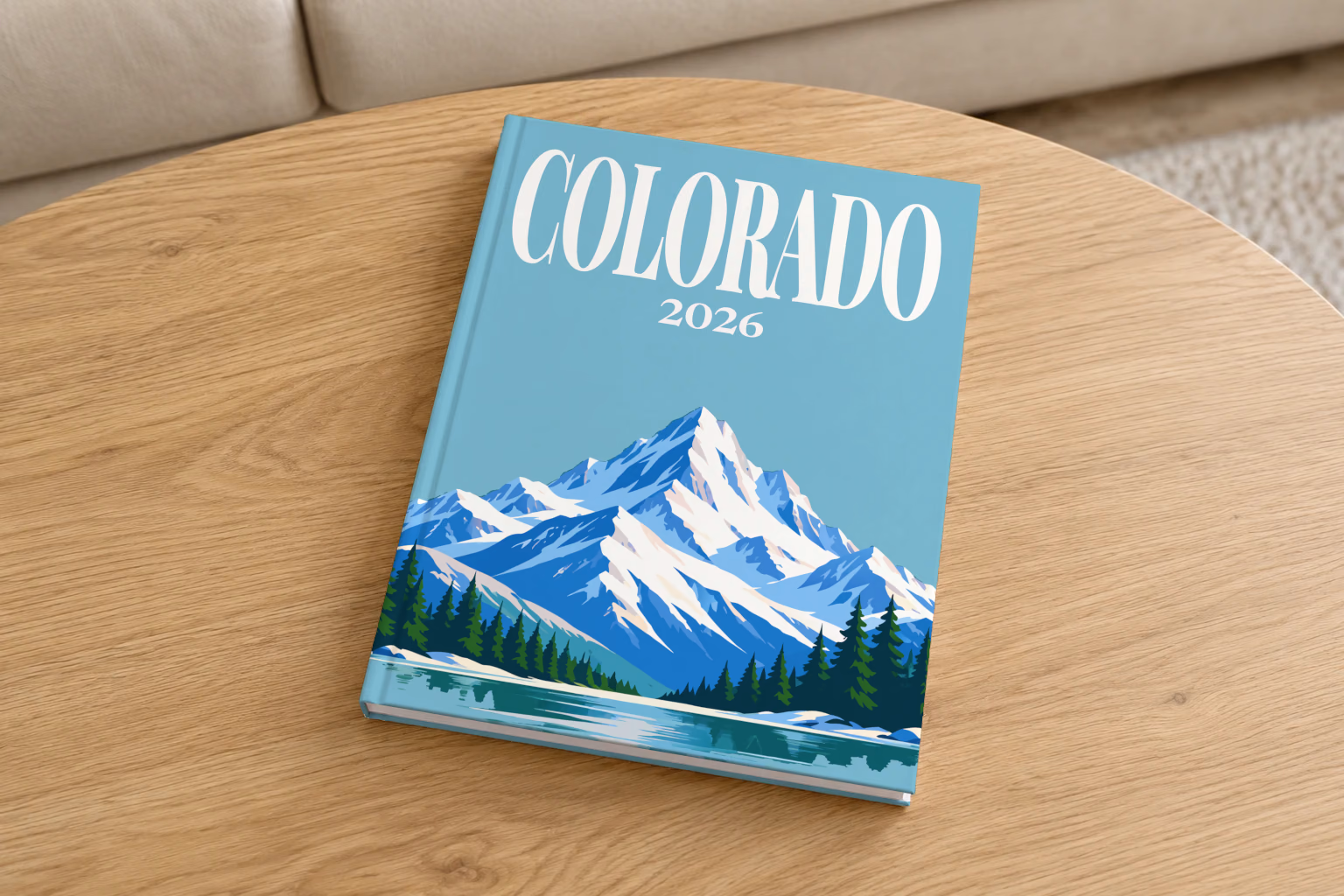 Colorado hardcover photo book with the illustrated cover of a snow-capped Rocky Mountain peak above a pine-lined lake on teal blue, on a round oak coffee table in a bright living room