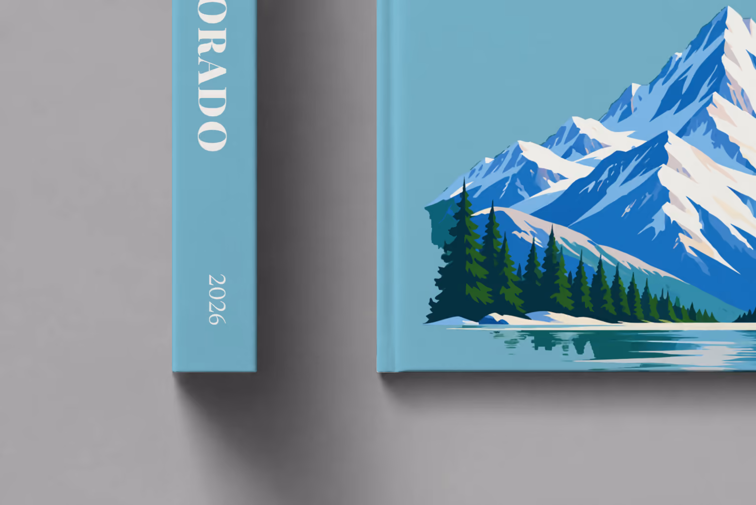 Colorado hardcover photo book with the illustrated cover of a snow-capped Rocky Mountain peak above a pine-lined lake on teal blue, shown close up beside its spine on a pale grey studio surface
