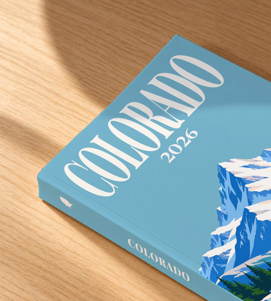 Colorado softcover photo book with the illustrated cover of a snow-capped Rocky Mountain peak above a pine-lined lake on teal blue, lying on an oak desk in dappled afternoon sunlight