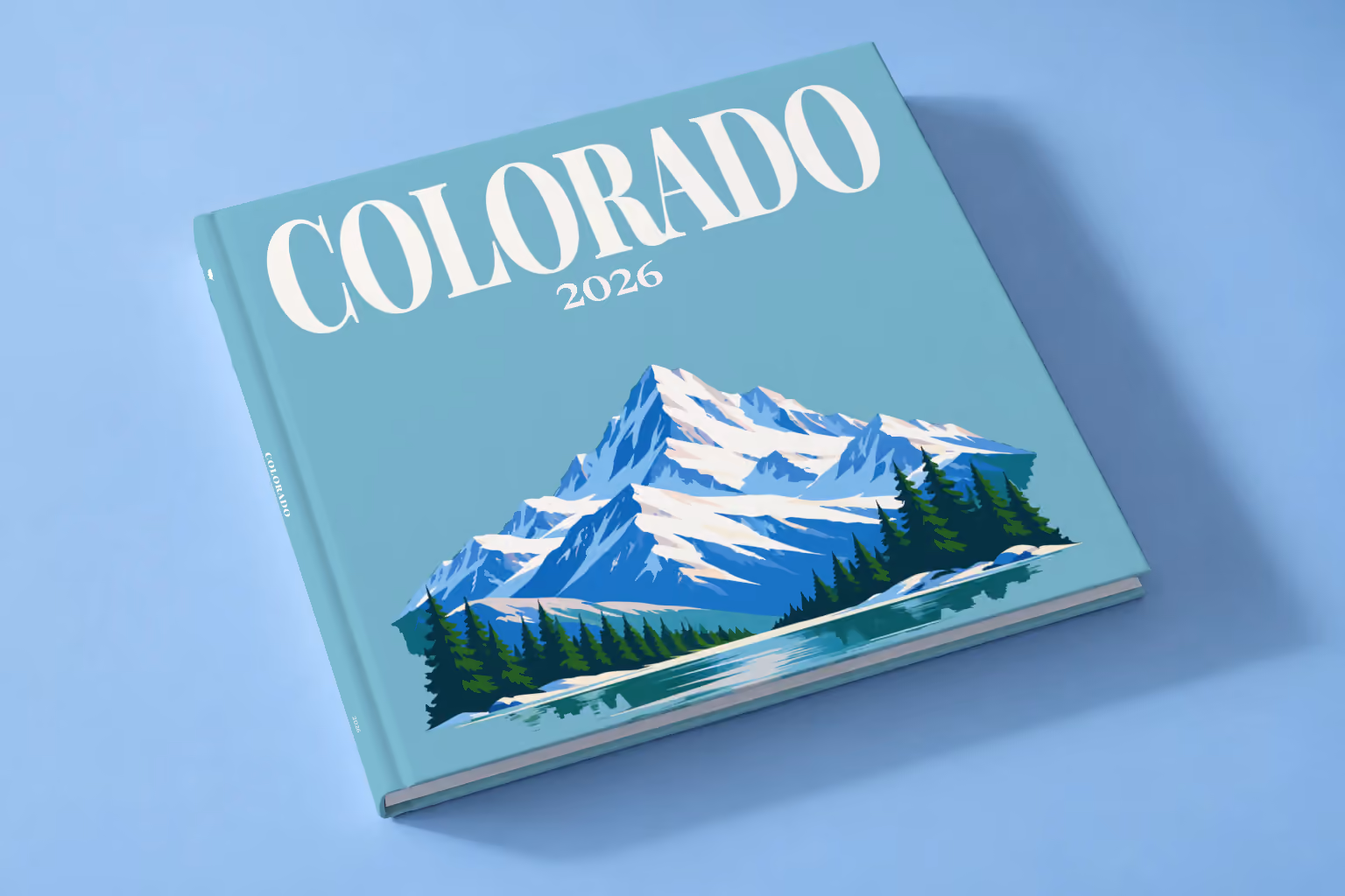 Colorado square hardcover photo book with the illustrated cover of a snow-capped Rocky Mountain peak above a pine-lined lake on teal blue, lying on a pale blue studio backdrop