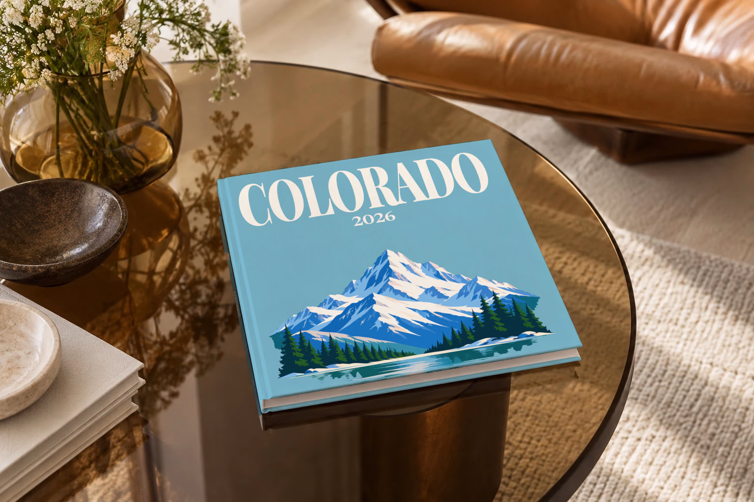 Colorado square hardcover photo book with the illustrated cover of a snow-capped Rocky Mountain peak above a pine-lined lake on teal blue, on a smoked glass coffee table beside a leather armchair in warm afternoon light