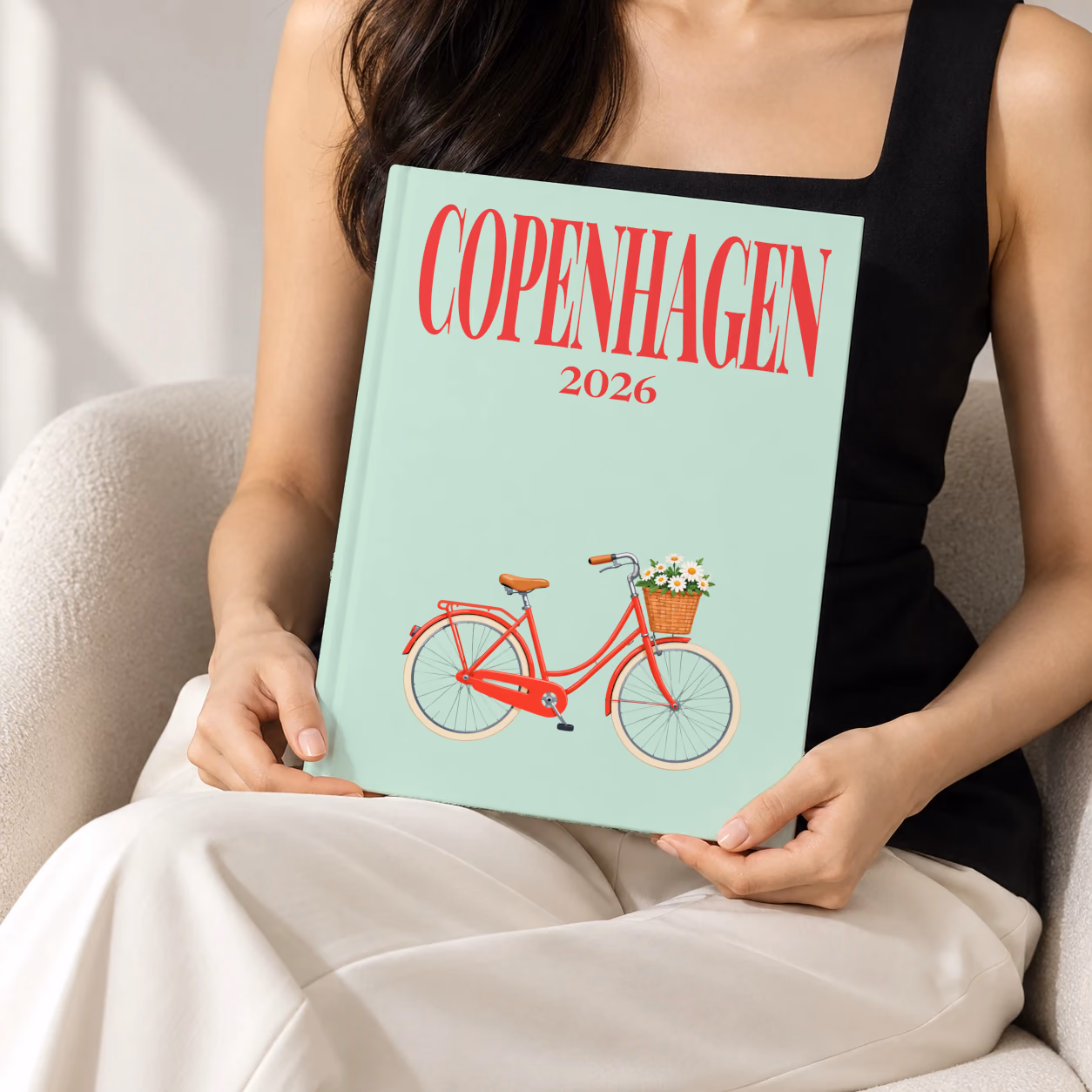 Denmark hardcover photo book with the Copenhagen illustrated cover of a red Copenhagen bicycle with a basket of daisies on mint green, held upright in two hands by a woman in a black top, sitting in a cream boucle armchair