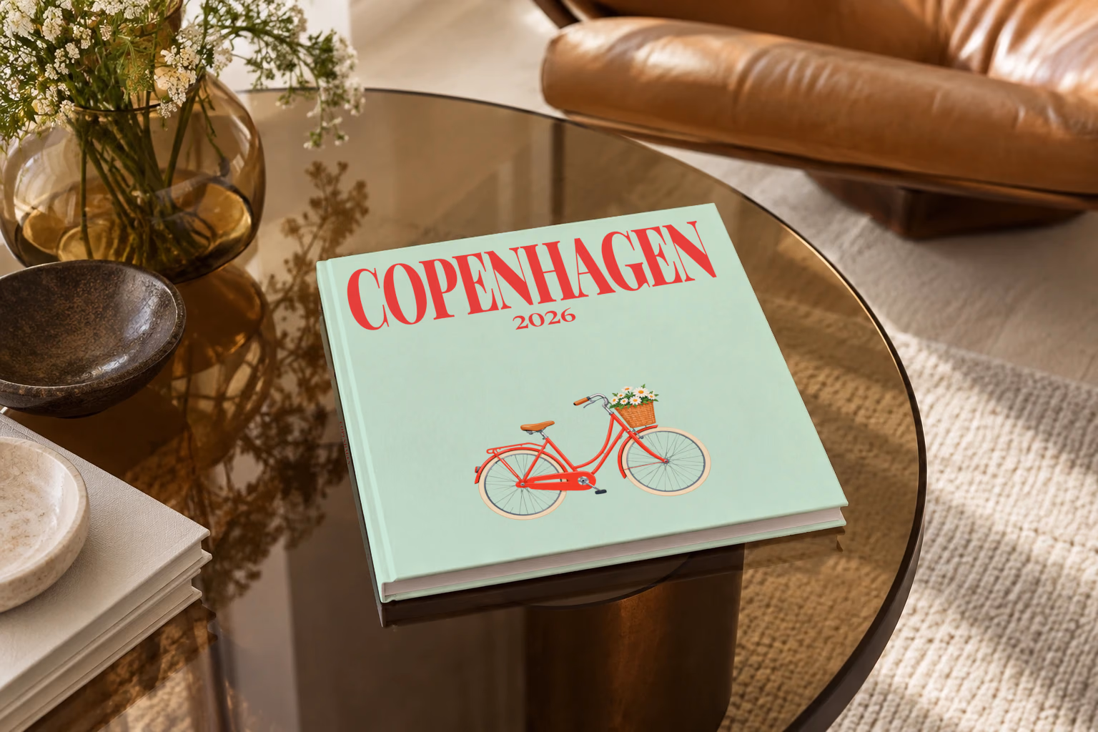 Denmark square hardcover photo book with the Copenhagen illustrated cover of a red Copenhagen bicycle with a basket of daisies on mint green, on a smoked glass coffee table beside a leather armchair in warm afternoon light