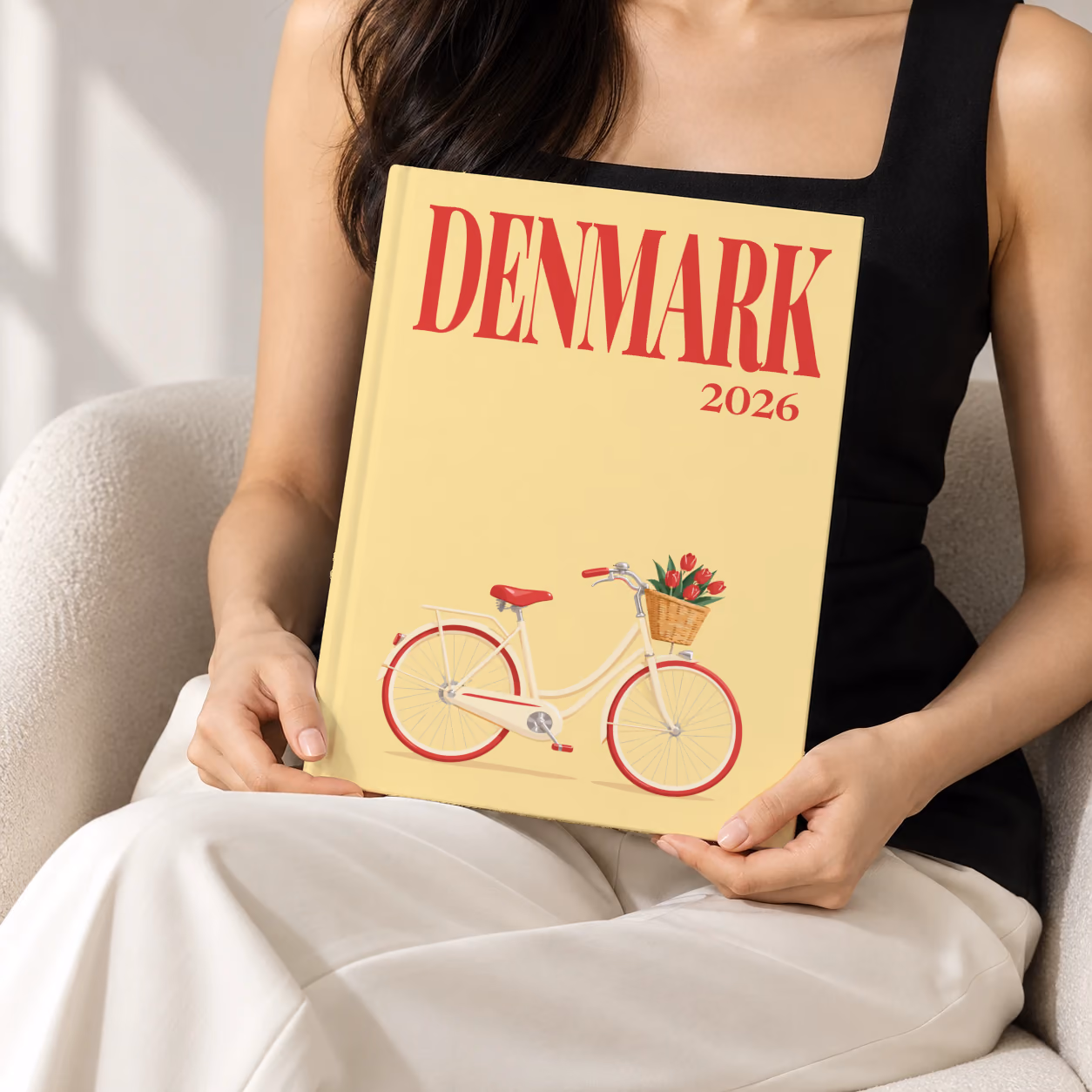 Denmark hardcover photo book with the illustrated cover of a Danish bicycle with a basket of red tulips on pale yellow, held upright in two hands by a woman in a black top, sitting in a cream boucle armchair