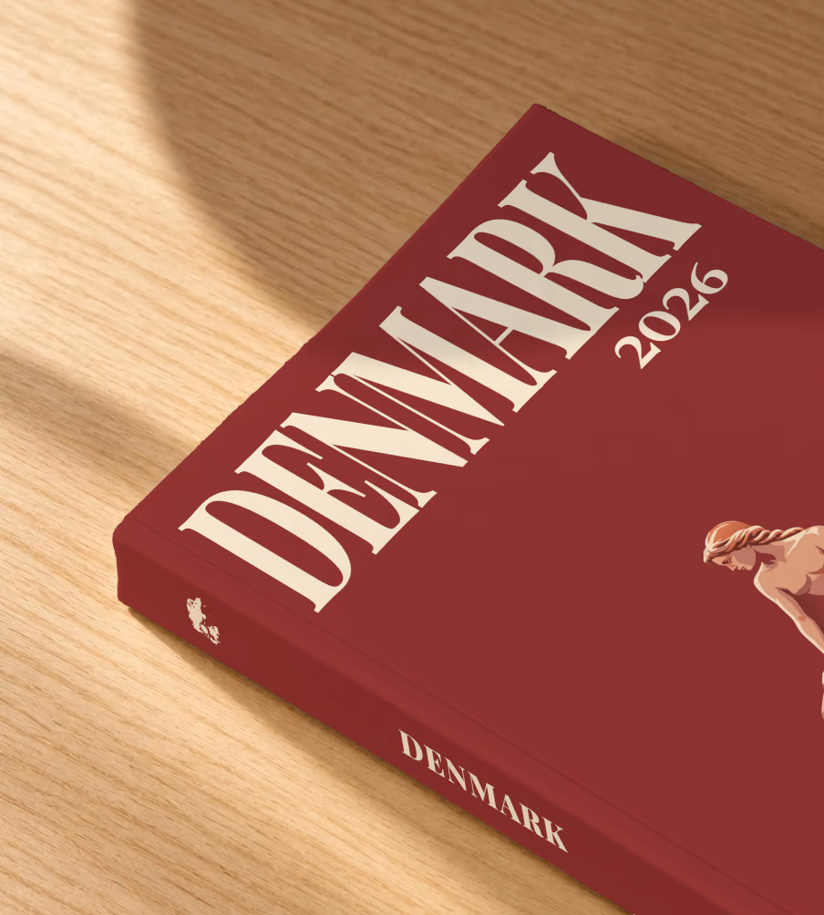 Denmark softcover photo book with the illustrated cover of the Little Mermaid statue on her rock in Copenhagen on burgundy, lying on an oak desk in dappled afternoon sunlight