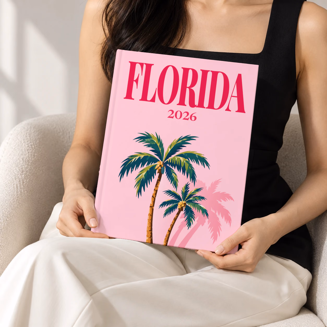 Florida hardcover photo book with the illustrated cover of Florida palm trees on pink, held upright in two hands by a woman in a black top, sitting in a cream boucle armchair